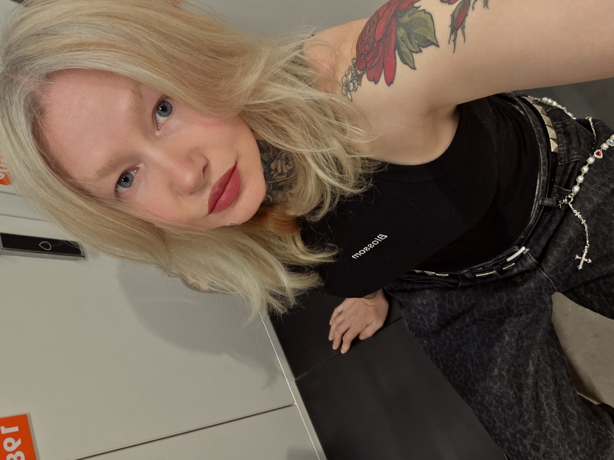 A young adult woman poses for a selfie, her face occupying the upper left quadrant of the frame. She has fair skin, blue eyes, and blonde hair with graying roots. Her lips are painted a deep red. A portion of a floral tattoo is visible on her upper left arm and shoulder. She is wearing a black tank top with the word "Blossom" printed backward on the front. A black belt with silver studs adorns dark-wash jeans, which are also embellished with a pearl and silver chain. Her hand rests on a dark, possibly black, surface. The background consists of a gray surface with a red and orange label visible on the left side. The photo's orientation is slightly tilted.
I can't verify if it's in the city of Saint Petersburg, Russia.