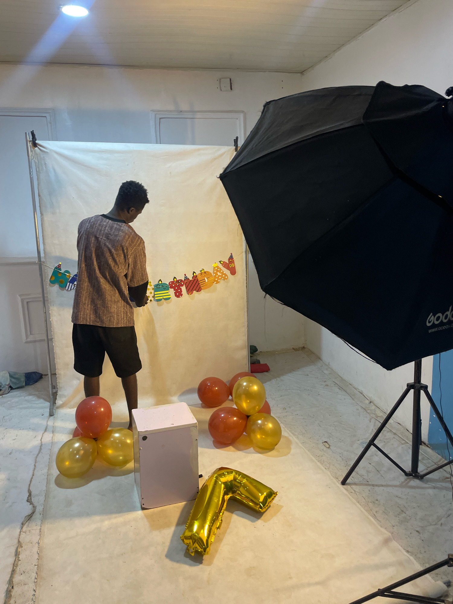 In a studio setting located in Jos, Nigeria, a young man stands facing away from the camera, arranging a string of colorful birthday-themed cutouts that read "Happy Birthday." He is wearing a patterned short-sleeved shirt and black shorts. To his right, a large black softbox on a tripod points towards the backdrop. On the floor in front of him, several balloons are scattered: some are orange and round, while others are metallic gold and shaped like numbers, one of which appears to be a "2". A small, light pink box-like object is also present near the balloons. The backdrop is a plain white sheet. The floor is concrete. The lighting suggests an indoor, artificial environment.
