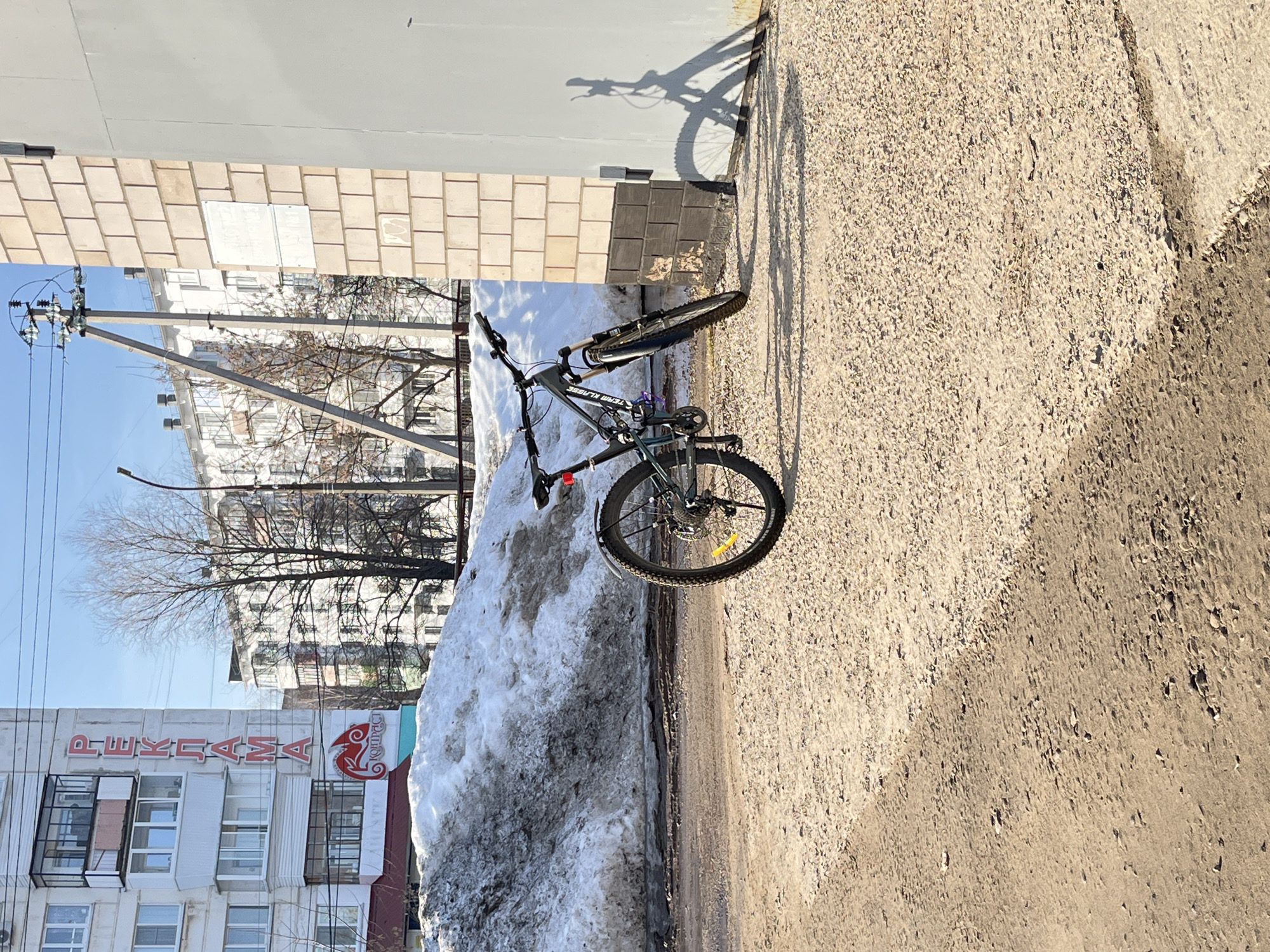 A bicycle is parked on an asphalt road next to a pile of dirty snow in Chaikovsky, Russia. The bike casts a shadow on the wall of a building to its right. In the background, buildings and trees are visible. Overhead power lines are to the left. A building with the word "PEKΛAMA" (advertisement) in red Cyrillic letters is partly visible.