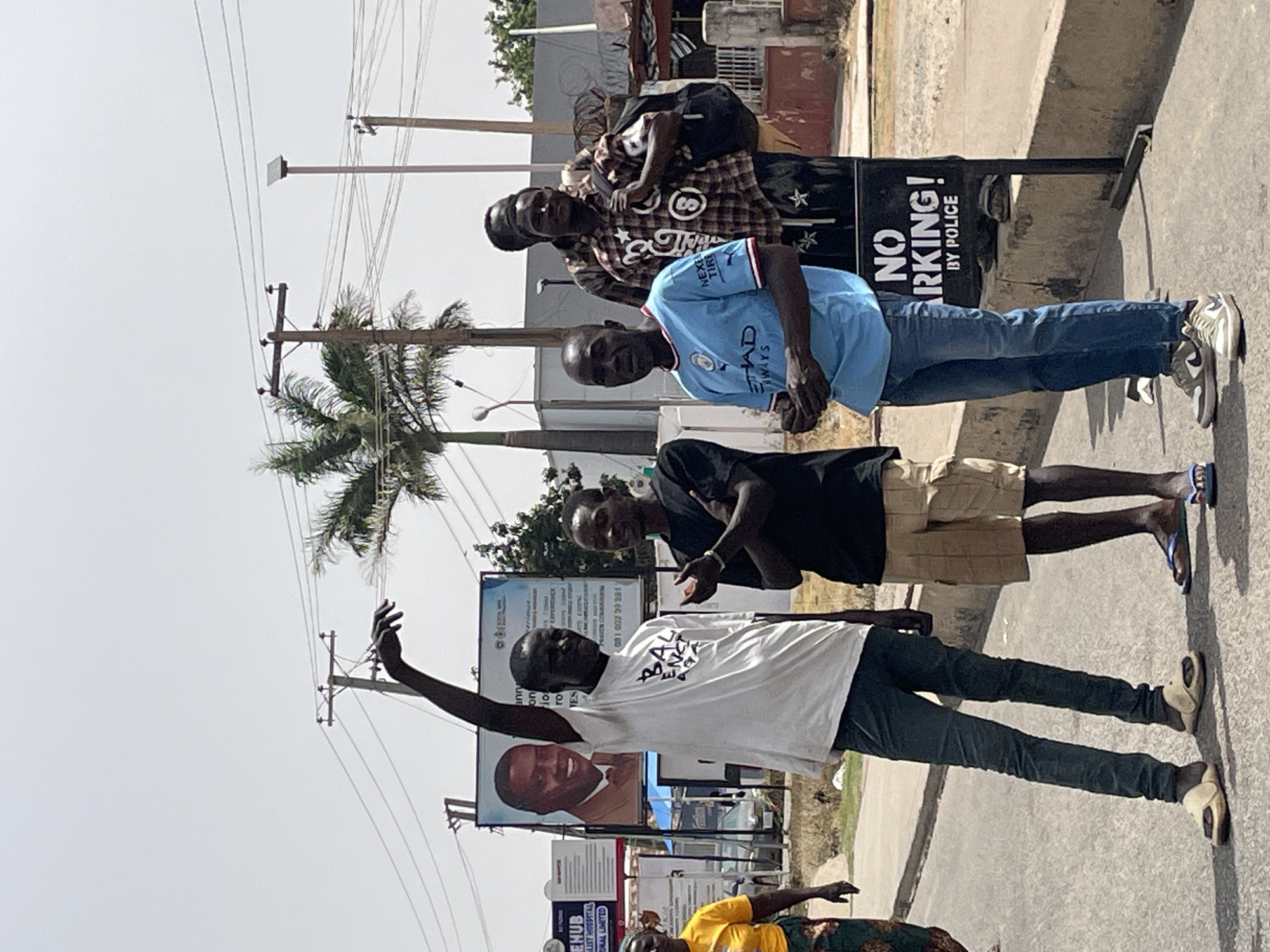 Four men stand together on a sidewalk beside a road, with power lines and a palm tree visible in the background. A "NO PARKING! BY POLICE" sign is next to one of the men. In the background, there is a billboard with a man's face and other signs, including one for ENUB LIST HOSPITAL, suggesting the location is in the city of Jos, Nigeria.