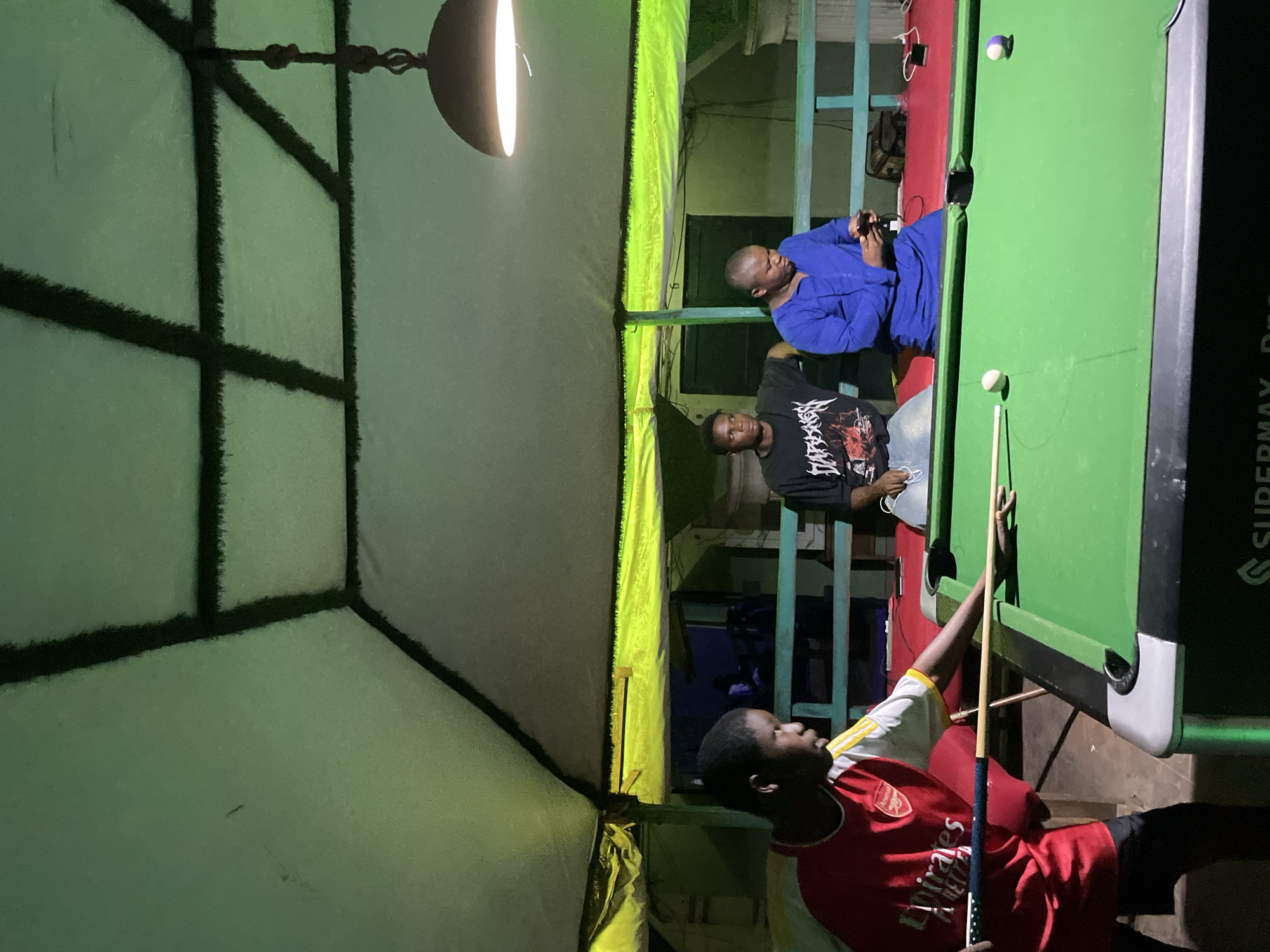 An indoor leisure space in Lokoja, Nigeria, is depicted under artificial illumination. Three dark-skinned males are present. A young male, wearing a red and white Arsenal football club jersey, is positioned over a green felt pool table, holding a cue and aiming at a white cue ball. A purple striped ball is also visible on the table. Behind the pool table, two adult males are seated on a low structure. One male, wearing a black t-shirt with a white graphic print, observes the pool game. The other male, dressed in a blue tunic, is holding and looking at a smartphone. The overhead structure features a light green segmented ceiling with dark support beams. Perimeter lighting in a yellow-green hue runs along the lower edge of the ceiling, and a single bright pendant lamp hangs centrally over the pool table. A teal-colored wooden railing is visible in the background, and a red floor covering is beneath the seated individuals. The pool table bears "SUPERMAX Pro" branding.