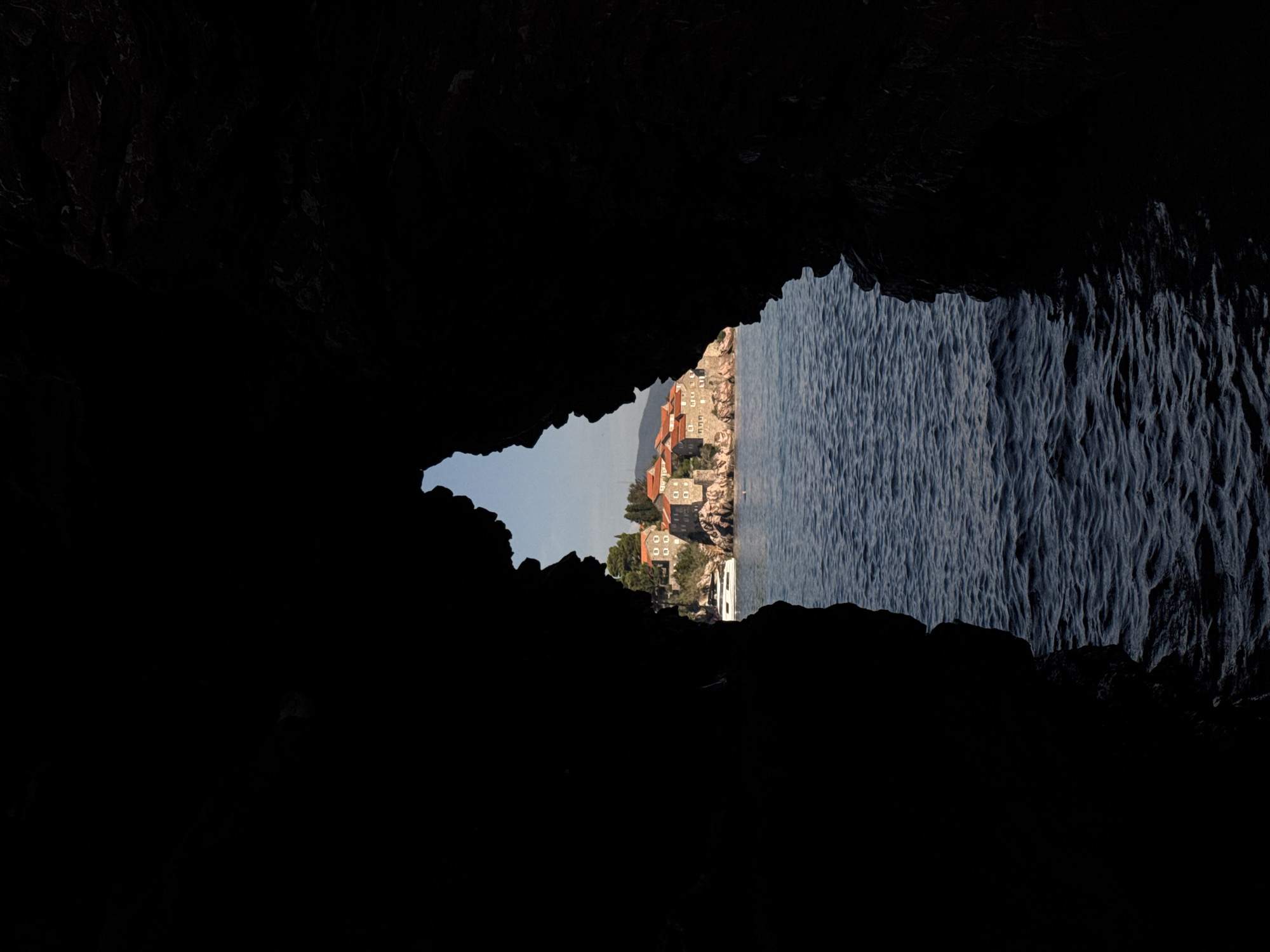 The image captures a view of Sveti Stefan, Montenegro, framed by the dark, silhouetted opening of a cave or rock formation. The foreground is dominated by the black mass of the cave entrance, creating a strong contrast with the scene beyond. Through the opening, a body of deep blue water with moderate waves is visible. On a rocky outcrop in the distance, several buildings with reddish-orange tile roofs are clustered together. These buildings appear to be constructed from stone. A white yacht is docked near the island. The sky above is a clear, light blue. The scene is likely captured from a boat or a water-level vantage point outside the cave.