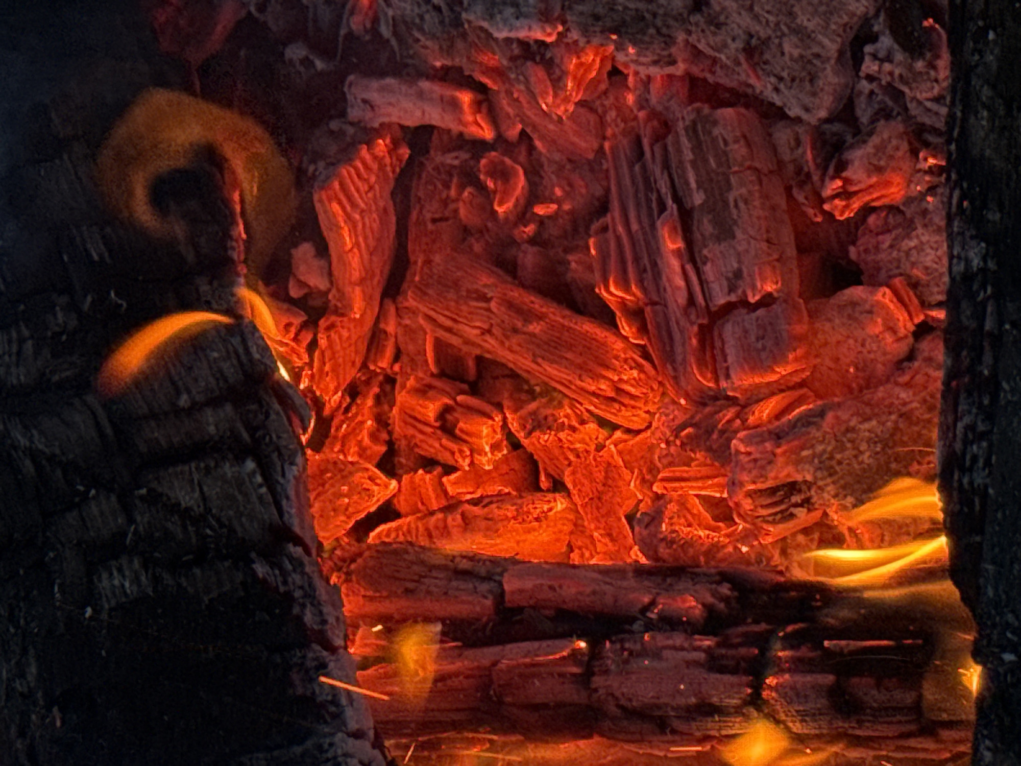 The image depicts a close-up view of a bonfire with glowing embers and flames. The dominant colors are red, orange, and black, highlighting the intense heat and texture of burning wood. The scene is set in Imelevo, Russia, and shows a close-up of burning logs, embers, and flames. There are no people or animals visible in the image. The environment appears to be outdoors, likely a natural setting given the context of a bonfire. The focus is on the dynamic and fiery nature of the combustion process, with the wood elements showing charred surfaces and the embers emitting a strong red glow.