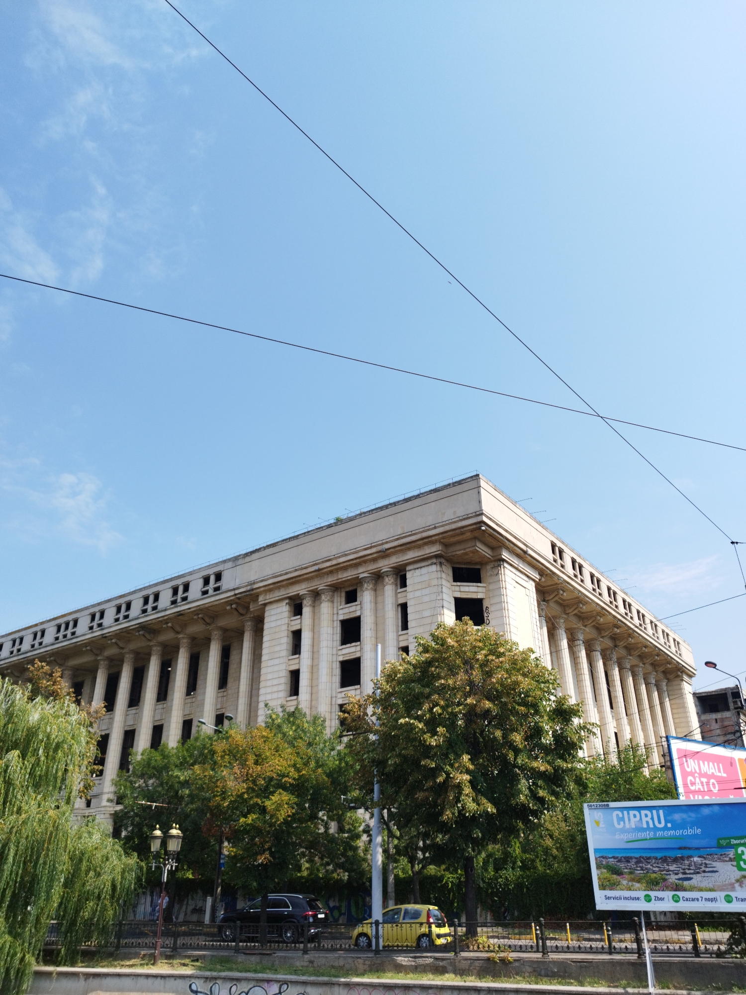 The image shows the Palace of the Parliament in Bucharest, Romania, a colossal building with a neoclassical facade featuring numerous columns and windows. The sky above is clear and blue with thin clouds and crisscrossing power lines. Trees are in the foreground, some with green leaves and others with autumnal colors, suggesting the season is late summer or early autumn. Several cars are visible on the street in front of the building, including a black SUV and a small yellow car. Two billboards are also present, one advertising "UN MALL CĂT O..." and another for "CIPRU. Experiențe memorabile," which is a travel advertisement for Cyprus. A street lamp stands near the road. The overall scene depicts a public space in a city with a mix of historical architecture, modern advertising, and everyday life.