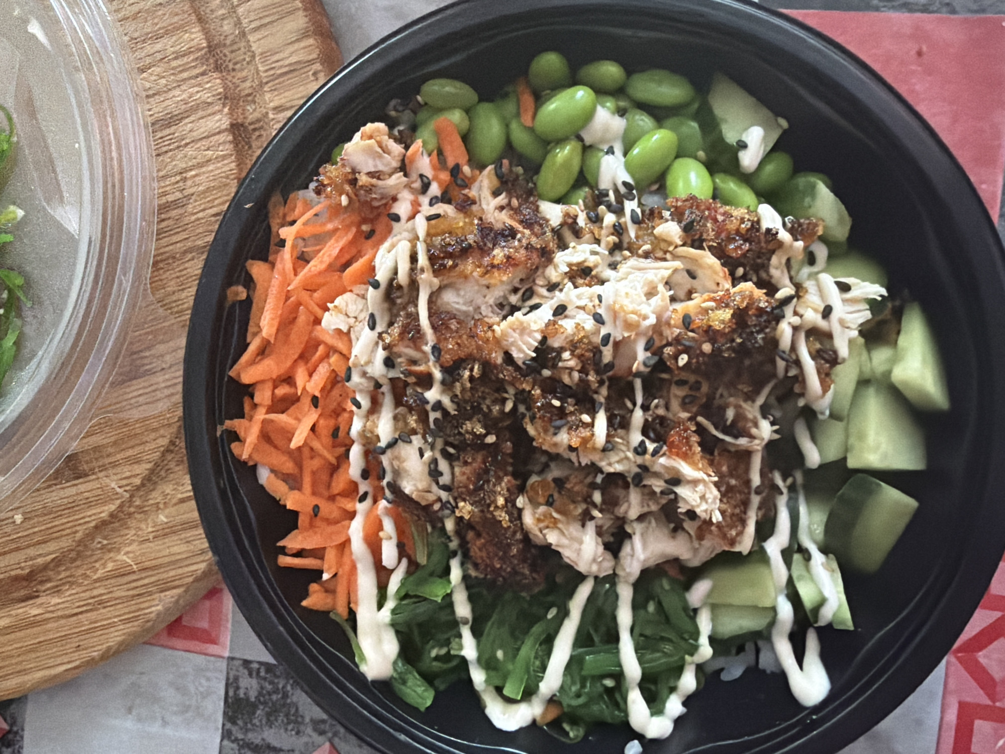 A top-down, close-up shot depicts a black bowl of prepared food, likely a poke bowl, set on a surface with a checkered pattern of red and grey/white squares. A wooden cutting board is partially visible to the left of the bowl.
The bowl contains the following ingredients arranged in sections: shredded carrots, cooked and shredded chicken with a crispy coating, edamame beans, and diced cucumbers. The bowl also contains green seaweed and a drizzle of creamy white sauce. Black sesame seeds are sprinkled on the chicken.
The location in the city of Plovdiv, Bulgaria is not discernible from the image.
