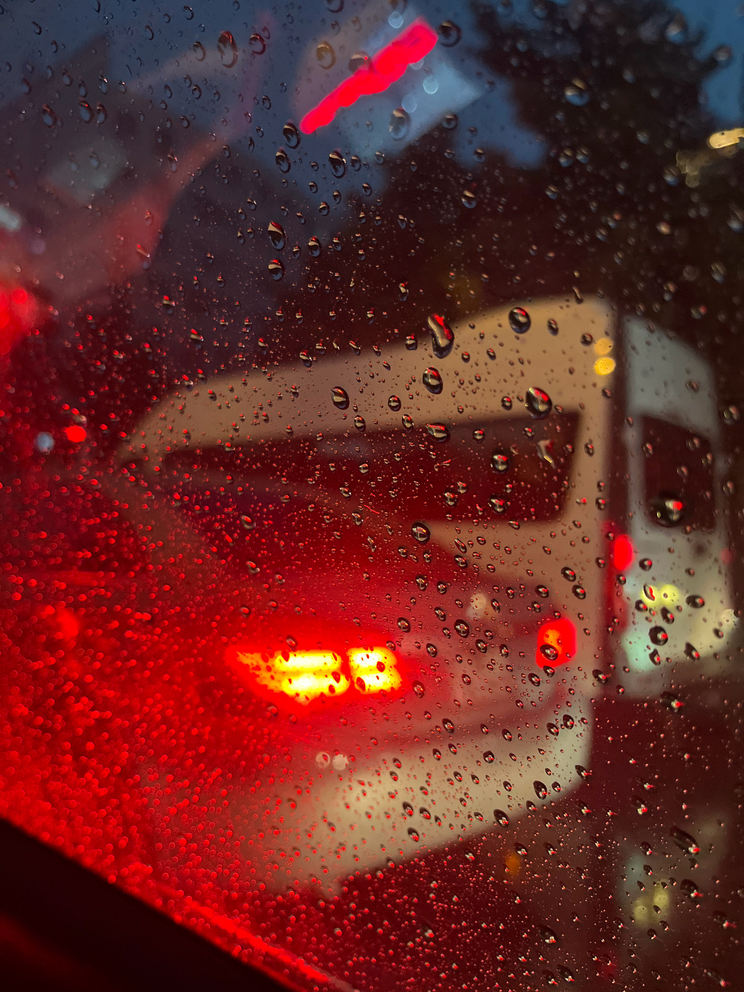 A photograph taken from inside a vehicle shows water droplets on the windshield, obscuring the view of traffic ahead. In the background, a white vehicle, possibly a bus or van, is visible with its red taillights illuminated. The scene appears to be a city street at dusk or night, with artificial lights from vehicles and possibly streetlights creating a blurred, colorful backdrop. The red light from the vehicle in front is particularly prominent, reflecting and refracting through the water droplets on the glass.