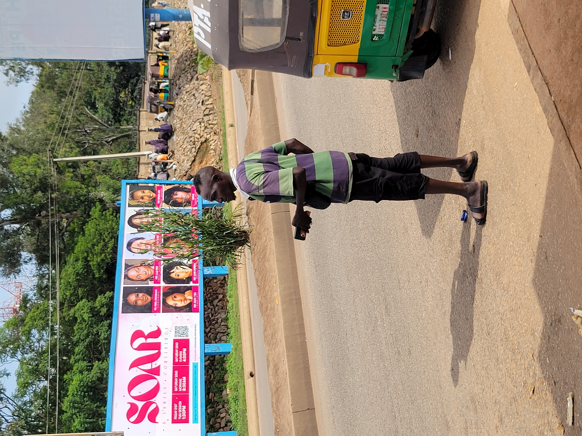 A man in a purple and green striped t-shirt and black shorts stands on a paved road, looking to his left. He is holding a dark object in his right hand and has a black sling bag across his chest. To his right is a yellow and green auto-rickshaw with a license plate that reads "LGT-732VM". In the background, a large billboard for "SOAR A LADIES CONFERENCE" is visible. The billboard features pictures of several women and event details. Beyond the billboard, there are trees and a stone embankment. The scene appears to be a street in Jos, Nigeria, with people and vehicles in the background suggesting a public area.