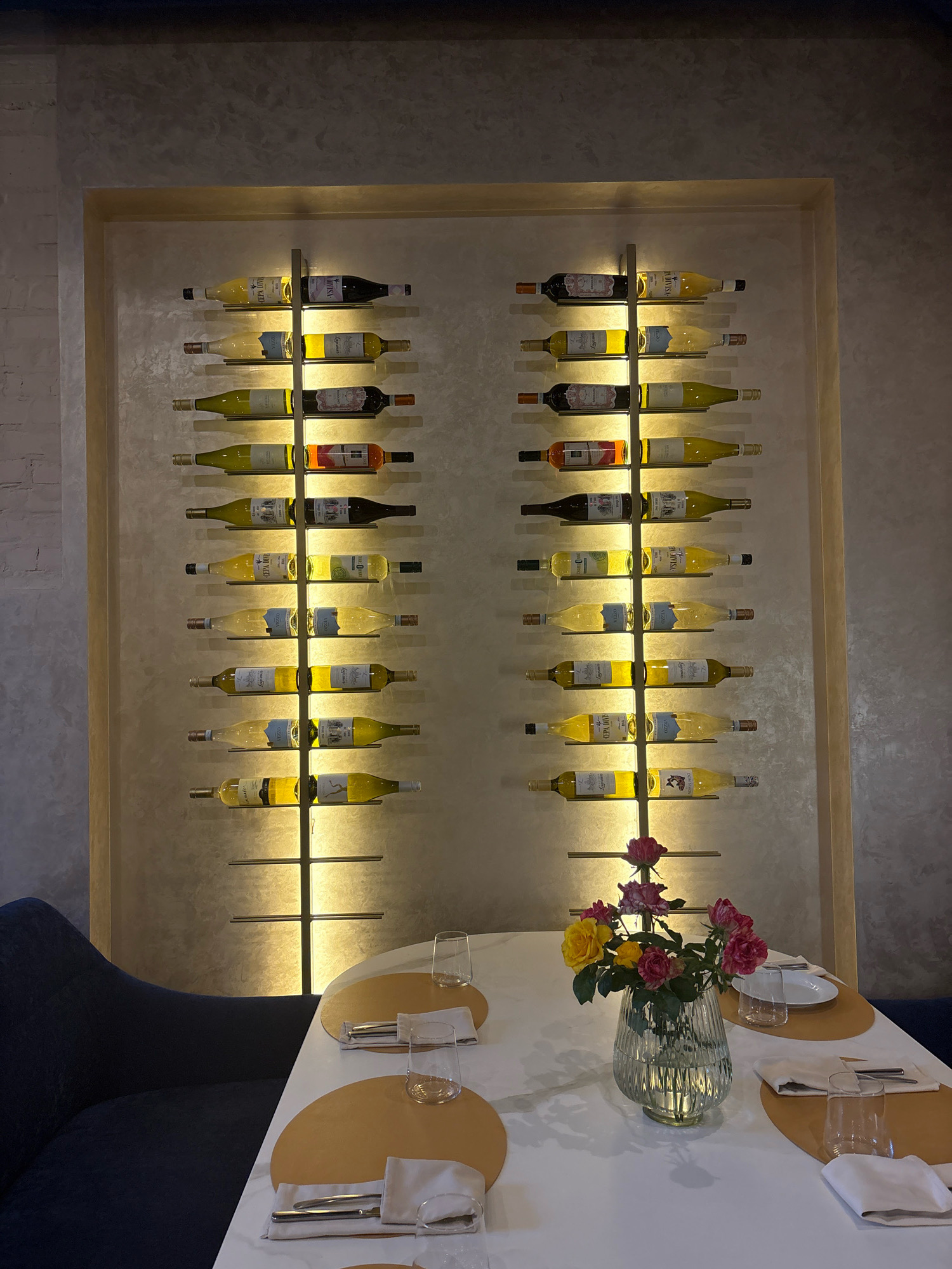 A dimly lit restaurant interior is depicted in Sofiivska Borshchahivka, Ukraine. A prominent wall-mounted wine rack displays numerous bottles of wine, illuminated by soft lighting. The rack is divided into two vertical columns, each holding eight bottles. On the right side of the rack, the top four bottles are clearly visible, with labels facing forward, revealing them to be mostly white wine. Below these, the next four bottles are also white wine, with labels visible. On the left side of the rack, the top four bottles are also clearly visible, with labels facing forward, revealing a mix of red and white wine. The next four bottles are white wine, with labels visible. In the foreground, a white table is set for dining with two place settings. Each setting includes a circular placemat, a white napkin, cutlery, a glass, and a plate. A vase filled with a bouquet of pink and yellow roses sits in the center of the table. To the left of the table, a dark blue upholstered chair is partially visible. The background features a textured wall with a subtle mottled pattern.
