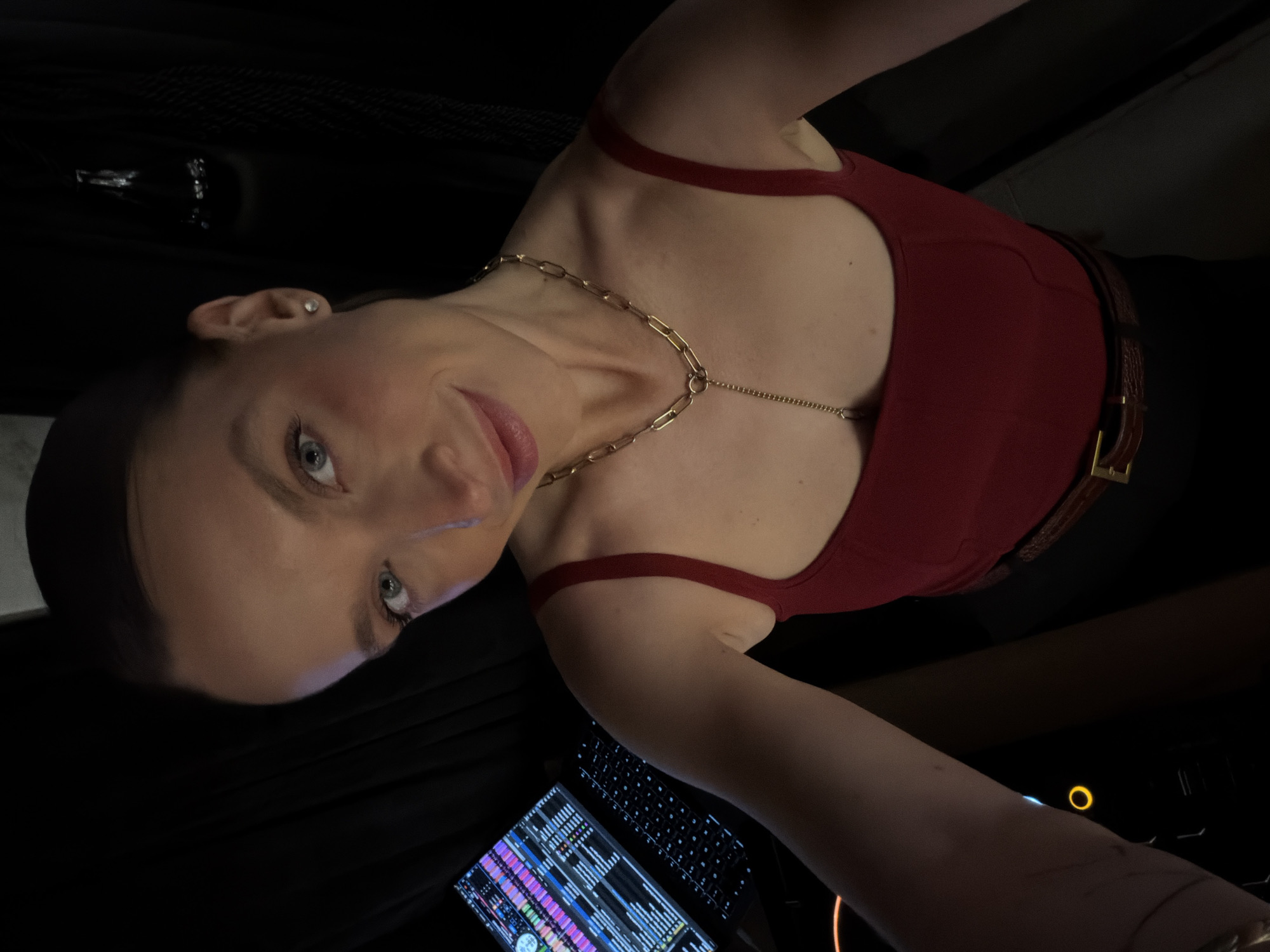 A woman is posing for a photograph indoors, likely in a dimly lit room, possibly in a venue such as a nightclub. She is wearing a dark red tank top, a brown belt, and a gold necklace. A laptop sits on the table near her. The laptop has a musical interface visible on the screen, indicating that music production or DJ activity may be taking place. Due to the lack of visible background information, the location in Budva, Montenegro, cannot be determined.