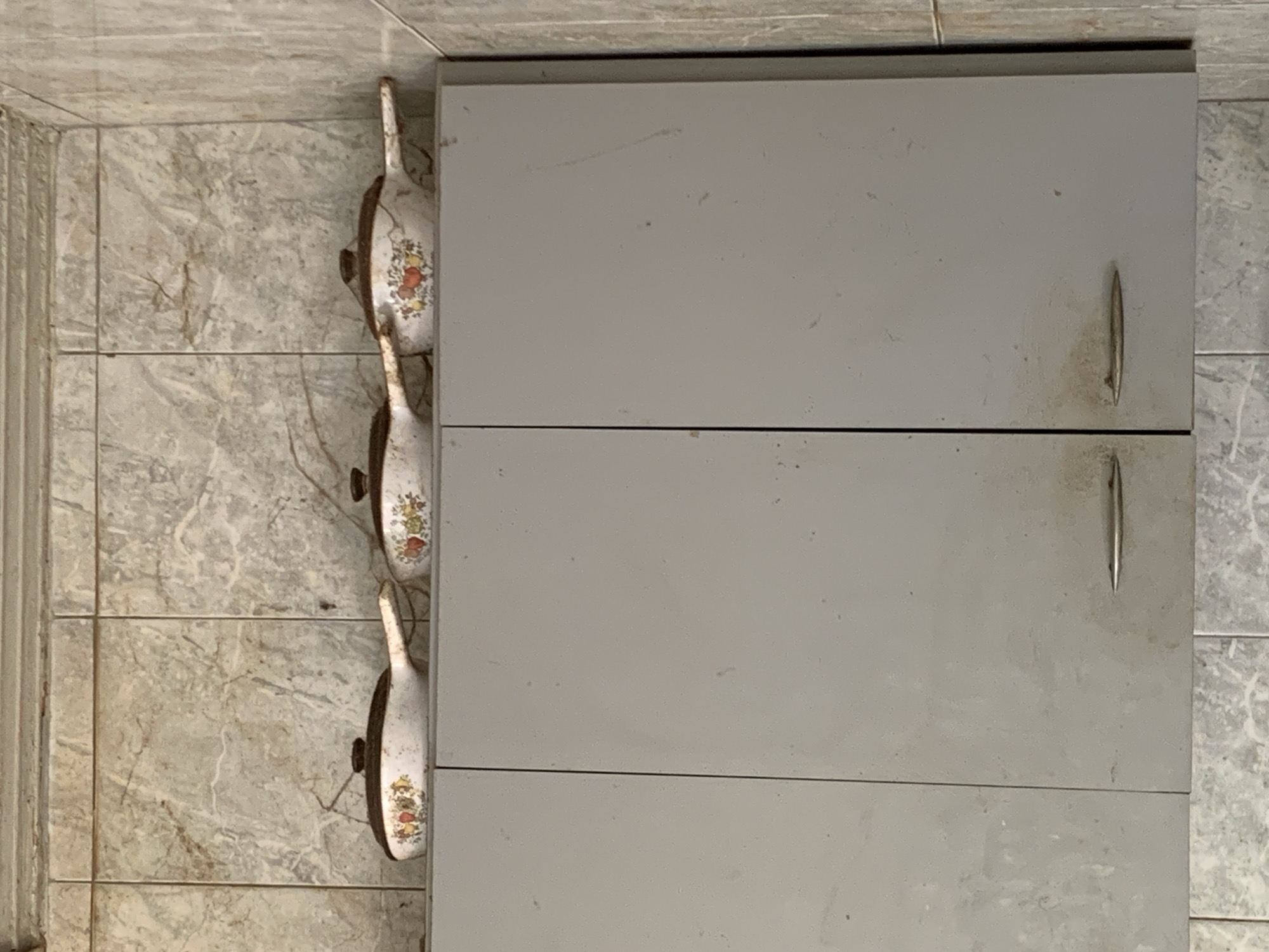The image shows a vertical shot of a gray metal cabinet mounted on a wall covered in patterned gray tiles. Three decorative, floral-patterned pans hang to the left of the cabinet. The scene appears to be indoors, possibly in a kitchen or storage area. The precise location within the city of Santa Ana, Venezuela is not ascertainable from the image.