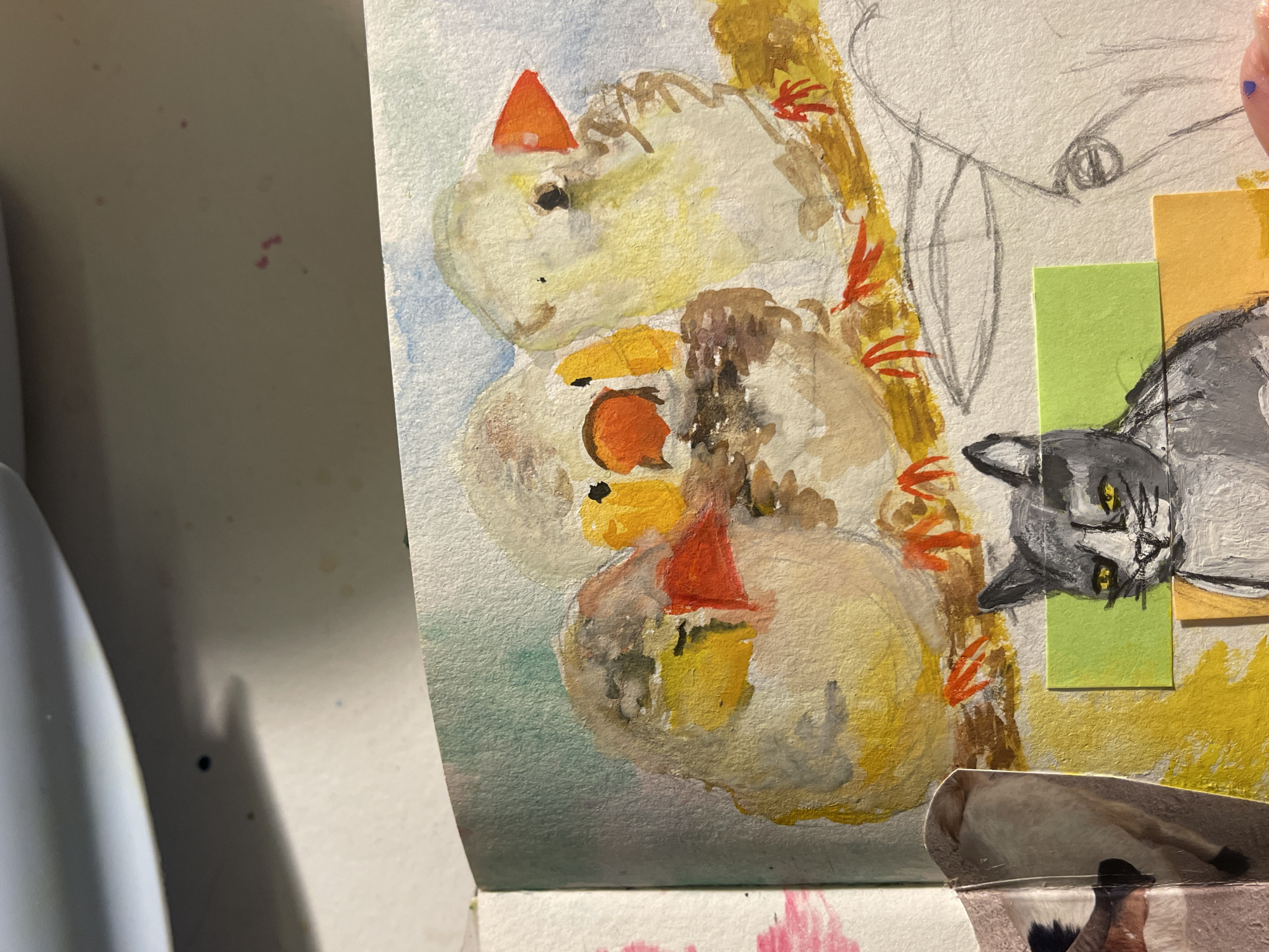 The image displays a close-up of a mixed-media artwork, likely in a sketchbook or art journal, featuring various drawn, painted, and collaged elements. The central focus is a stack of three rounded, light-yellow figures, resembling abstract snowmen or creatures, each adorned with a bright orange cone-shaped hat. These figures are rendered with watercolor or thinned paint, showing washes of yellow, brown, and hints of blue in the background.
To the right of these figures, a gray cat's head with striking yellow eyes is painted on a separate piece of paper and appears to be collaged onto the artwork. Below this, another collaged element shows a portion of a dog's leg and paw. In the upper right section, a pencil sketch of an eye and other lines is visible on the bare paper, suggesting an unfinished drawing or preliminary sketch.
The artwork incorporates various textures: the slight roughness of watercolor paper, the matte finish of paint, and the smooth appearance of the collaged paper cutouts. Small, vibrant orange and red brushstrokes, possibly depicting leaves or flames, are visible around the central stack of figures. The overall scene suggests an artistic workspace, with faint pink paint splatters visible on the white surface to the left and below the main artwork. A light-colored, curved object, possibly a pen or the edge of a container, is partially visible on the far left. No explicit text is discernible in the image.