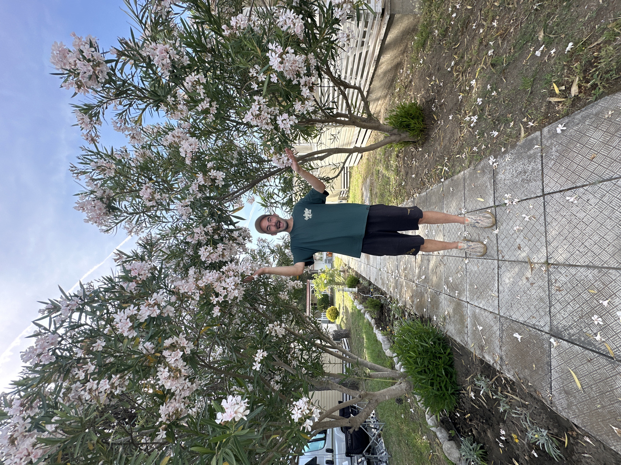A man stands with his arms outstretched near a flowering tree. He is wearing a green t-shirt, black shorts, and a hat. A paved path leads away from him, lined with flowering trees and greenery. White flower petals are scattered on the ground. A camper van is partially visible behind the tree. The scene is located in Donji Štoj, Montenegro.