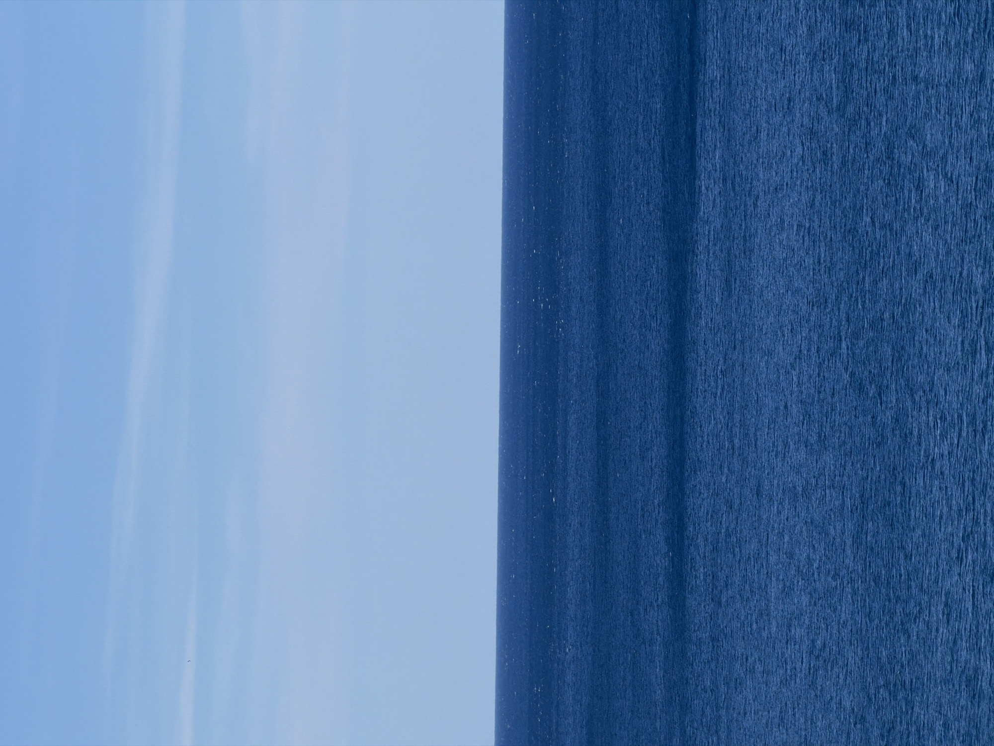 The image depicts a view of the Adriatic Sea meeting the sky in Mirište, Montenegro. The left side of the image shows a light blue sky with faint cloud streaks. The right side of the image shows the dark blue, textured surface of the sea. The boundary between the sky and the sea appears to be a relatively smooth horizon line, with a slightly darker strip of water near the horizon. The photograph is oriented vertically. There are no visible people or objects within the scene other than the sky and the sea.