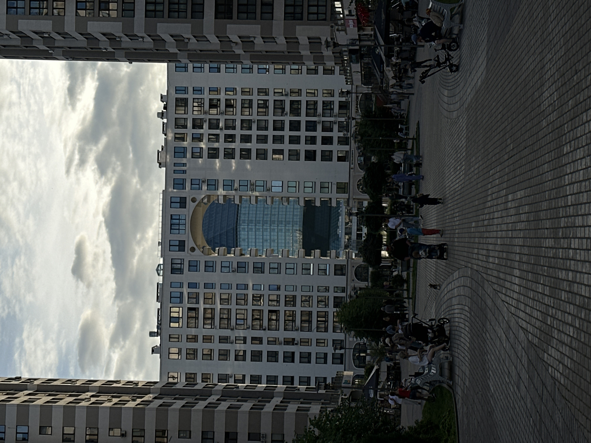The media file depicts an outdoor scene featuring modern architecture and people. Two large buildings, likely apartment complexes or office buildings, dominate the background. The building on the right has a distinctive rounded facade with a central section clad in glass, reflecting the cloudy sky. The building on the left is a more rectangular structure with repetitive window patterns.
In the foreground, a paved area with a curved design suggests a public space, possibly a plaza or promenade. Several people are visible, engaged in various activities. Some are walking, while others appear to be sitting on benches or pushing strollers. Several electric scooters and a bicycle are also present, indicating a modern urban environment where such personal transportation is common.
The time of day appears to be late afternoon or early evening, given the lighting conditions and the soft, diffused sunlight filtering through the clouds. The weather is overcast, with significant cloud cover. The overall mood of the scene is relaxed and everyday, with people enjoying their time outdoors. There are no discernible emotions conveyed by the individuals, as they are mostly captured from a distance. No visible text is present in the image.