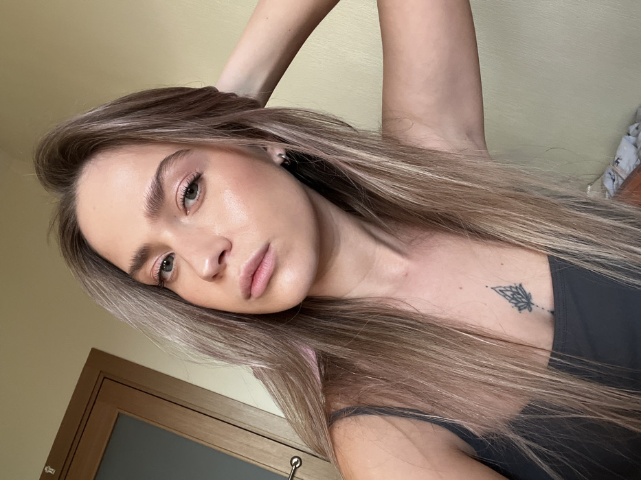 A female individual is depicted in a close-up portrait, facing the camera directly in an indoor setting within Saint Petersburg, Russia. She has long, straight to slightly wavy light brown hair with blonde highlights, green eyes, and wears light makeup. She is dressed in a dark gray strappy top. A small, black outlined tattoo, appearing as a stylized geometric floral design, is visible on her upper sternum. Her left hand is raised with fingers touching her hair on the viewer's right side of her head. The background consists of a plain light brown wall, and a wooden door with a frosted glass panel and a metallic handle is partially visible to the left. A small portion of patterned fabric is discernible in the lower right.