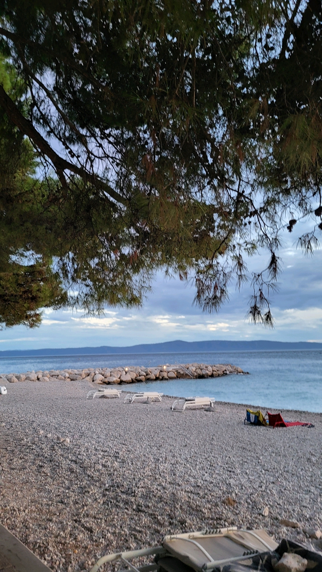 The image depicts a pebbly beach on the coast of Općina Tučepi, Croatia, under a cloudy sky. In the foreground, several empty lounge chairs are scattered across the beach, with a couple of brightly colored beach bags and towels lying near the water's edge. A stone jetty extends into the turquoise sea, with faint lights visible on it. Overhead, the branches of a large pine tree cast shadows, framing the scene. The overall atmosphere is tranquil and serene, suggesting either early morning or late evening.