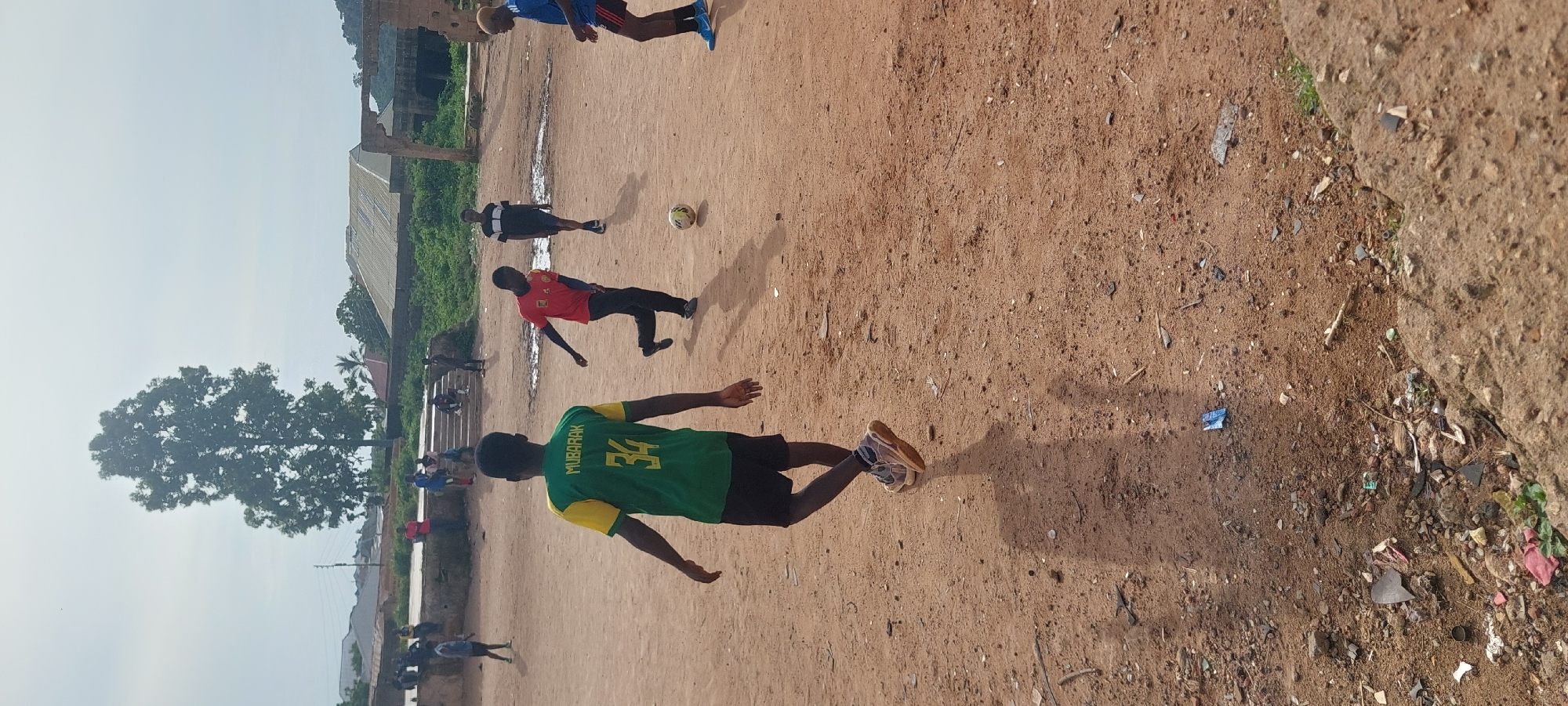 In the city of Bukuru, Nigeria, a group of young males are engaged in a game of football on a dirt field under an overcast sky. The field is uneven and scattered with debris. In the foreground, a young male wearing a green and yellow jersey with "MUBARAK" and the number "34" printed on the back is walking away from the camera, his shadow stretching across the dirt. To his right, another young male in a red t-shirt and dark pants is kicking the football. Further back, other individuals are visible, some watching the game and others participating. The background reveals a rural or semi-urban environment with buildings, trees, and lush vegetation. The overall scene depicts a casual football match in an undeveloped area.