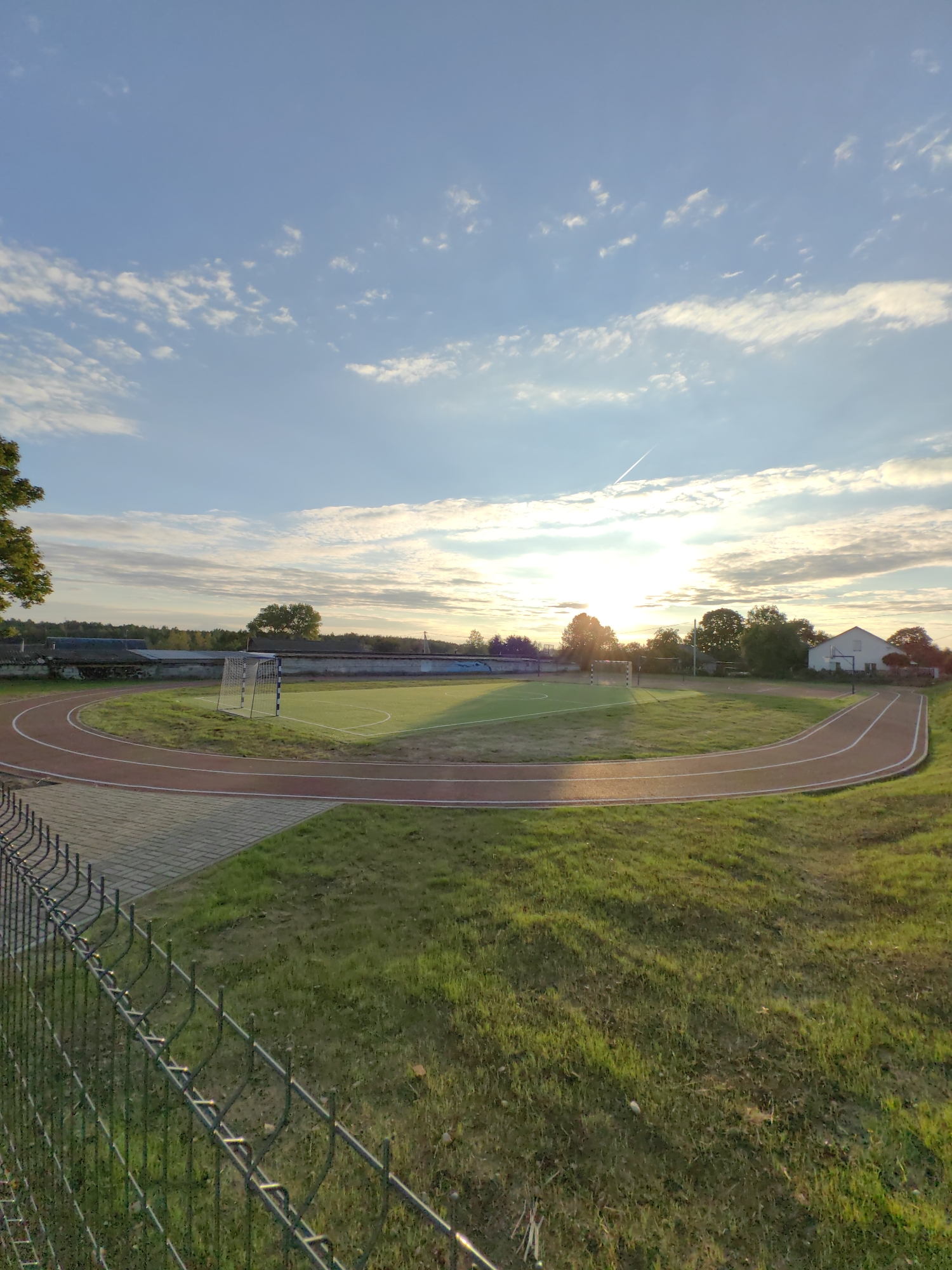 The image depicts a sunset at an outdoor sports facility in Myadzyel, Belarus. In the foreground, a chain-link fence borders a grassy area. Beyond the fence, a red running track curves around a green field, which appears to be a soccer field with a goalpost. The sun is low in the sky, casting long shadows and illuminating the clouds. Several trees and buildings are visible in the background, along with a wall surrounding the sports complex. The sky is blue with scattered white clouds and a faint contrail from an airplane.