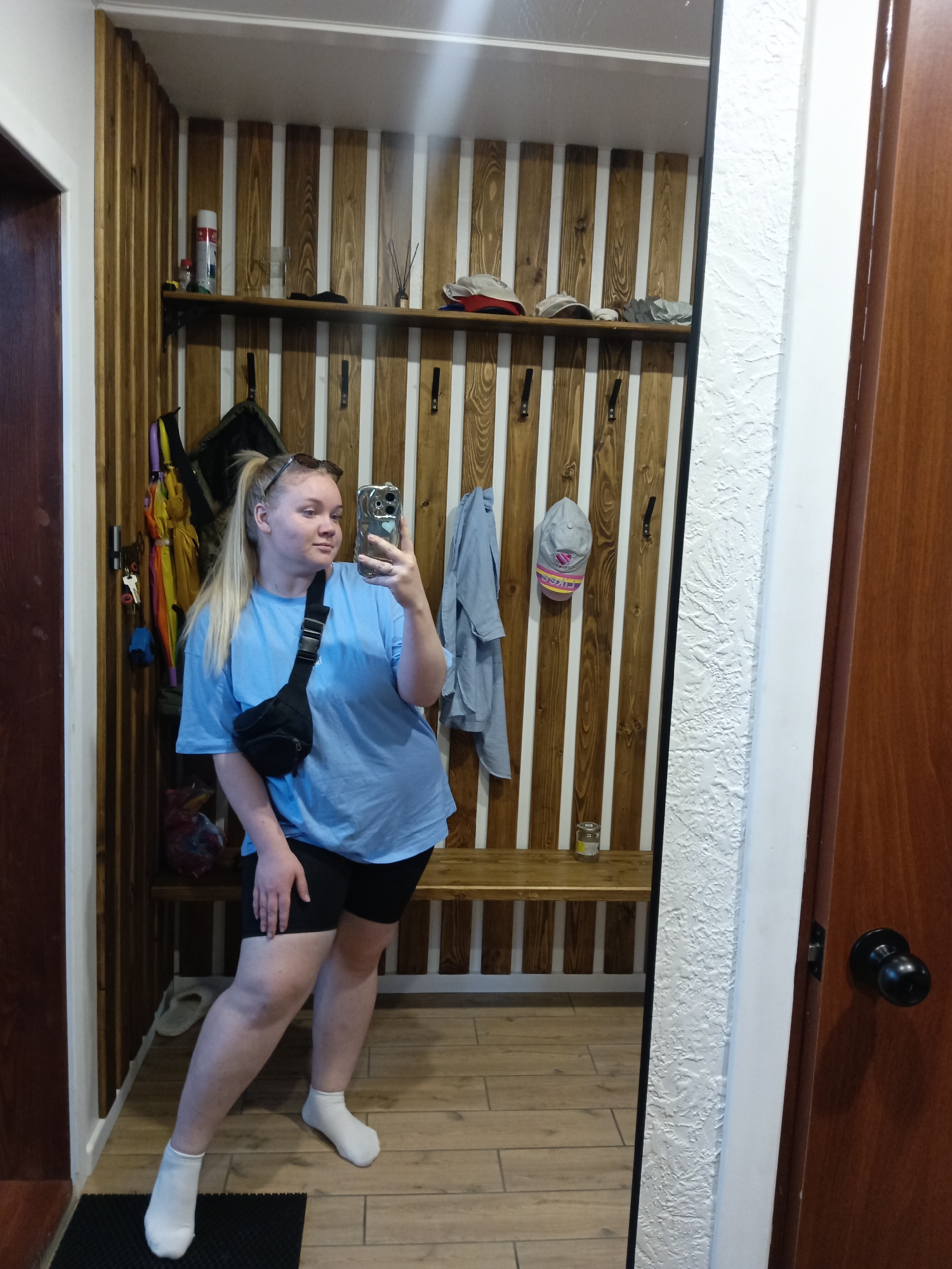 A young woman is taking a selfie in what appears to be an entryway or hallway. She has blonde hair pulled back in a ponytail and is wearing a light blue t-shirt, black biker shorts, and white socks. A black fanny pack is slung across her chest.
Behind her is a built-in wooden storage area with alternating dark wood planks and white vertical stripes. On the shelf above, there are various items including bottles, hats, and decorative sticks. Coat hooks are present, holding items like an umbrella, a bag, and a blue shirt. A wooden bench runs along the bottom of the storage area.
To the right is a wooden door with a black doorknob. The floor is tiled in a light brown color.
The location is Фоки, Russia.