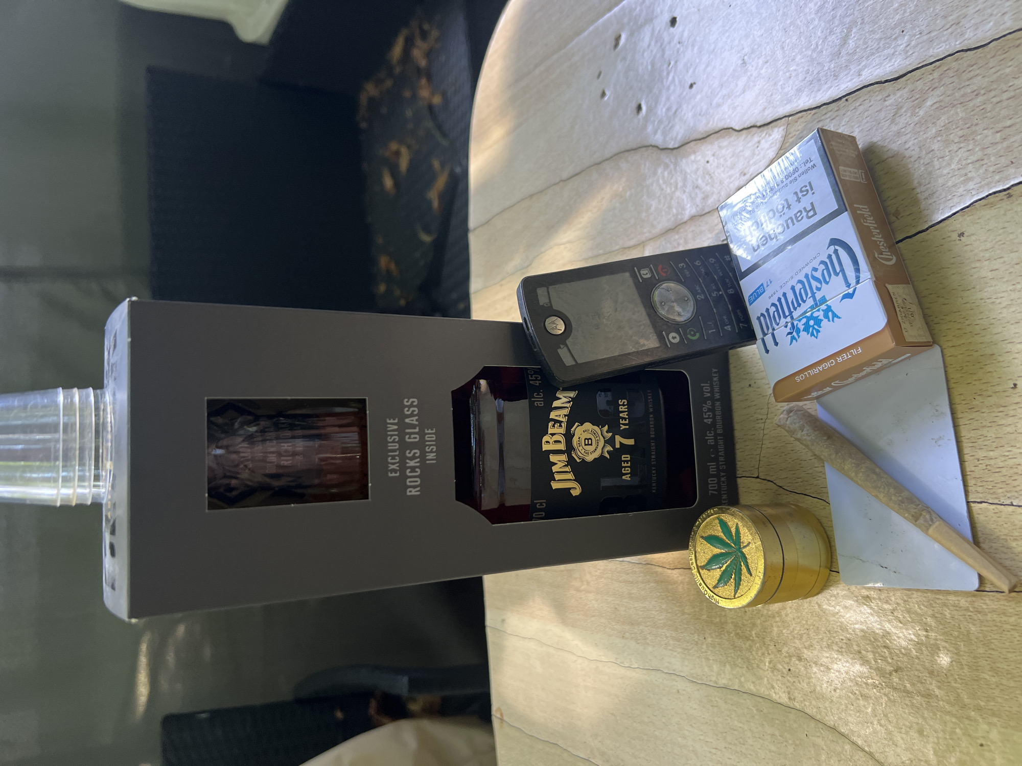 The image depicts a tabletop setting outdoors, likely in a backyard or on a patio. The scene includes a bottle of Jim Beam whiskey with an "exclusive rocks glass inside" presentation box, a pack of Chesterfield cigarettes, a metal grinder with a cannabis leaf design, a rolled cigarette (joint), and an old-style mobile phone. The items are arranged on a wooden surface that appears weathered, with visible grain and some small imperfections. In the background, a dark, blurred area suggests outdoor furniture, possibly a couch or chairs, and some fallen leaves. The lighting indicates it is daytime, with some shadows cast on the tabletop. There are no people or identifiable landmarks that would place the scene specifically in Wuppertal, Germany.