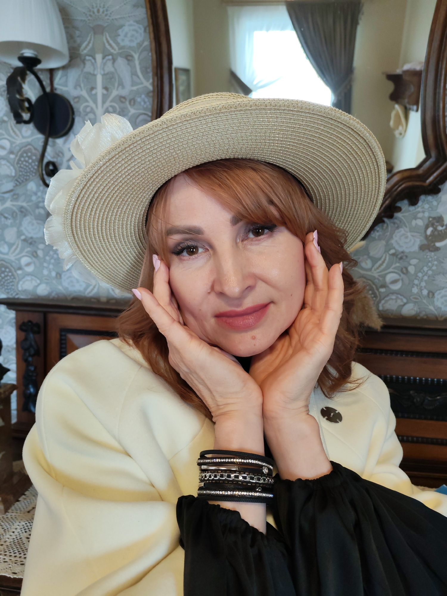 A woman with red hair is wearing a tan sunhat with a large white flower on the side. Her hands are raised, framing her face. She is wearing a cream-colored jacket over a black blouse and several black bracelets on her left wrist. The background features patterned wallpaper, dark wood furniture, a decorative sconce, and a mirror reflecting a window with curtains. The location is Sarapul, Russia.