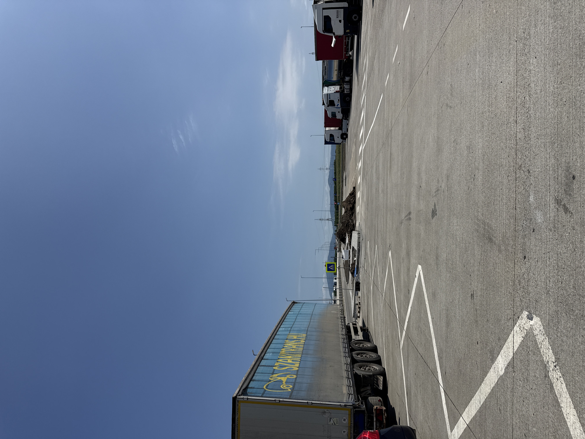 The image depicts a wide, paved area, likely a truck stop or parking lot, under a clear blue sky with a few wispy clouds. In the foreground, a large semi-trailer truck with a blue tarp and yellow lettering that reads "SZAKITRANS.HU" is parked on the left. The truck has visible wheels and a tandem axle. White painted lines on the pavement indicate parking spaces, with a zig-zagging line prominent in the lower right quadrant.
In the middle ground, several other trucks are parked in a row. These include a white truck with a red trailer, a white truck with a tank trailer, and another white truck with a red trailer. To the left of these trucks, there is a pile of dirt or construction debris. A yellow pedestrian crossing sign is visible on the right side of the image, near a road or entrance. In the distance, power lines and hills can be seen.
The location is identified as Sanpetru, Romania. The scene appears to be a functional commercial area, possibly a rest stop or a depot for freight transportation. There are no people visible in the image. The overall impression is one of an open, industrial outdoor space on a sunny day.