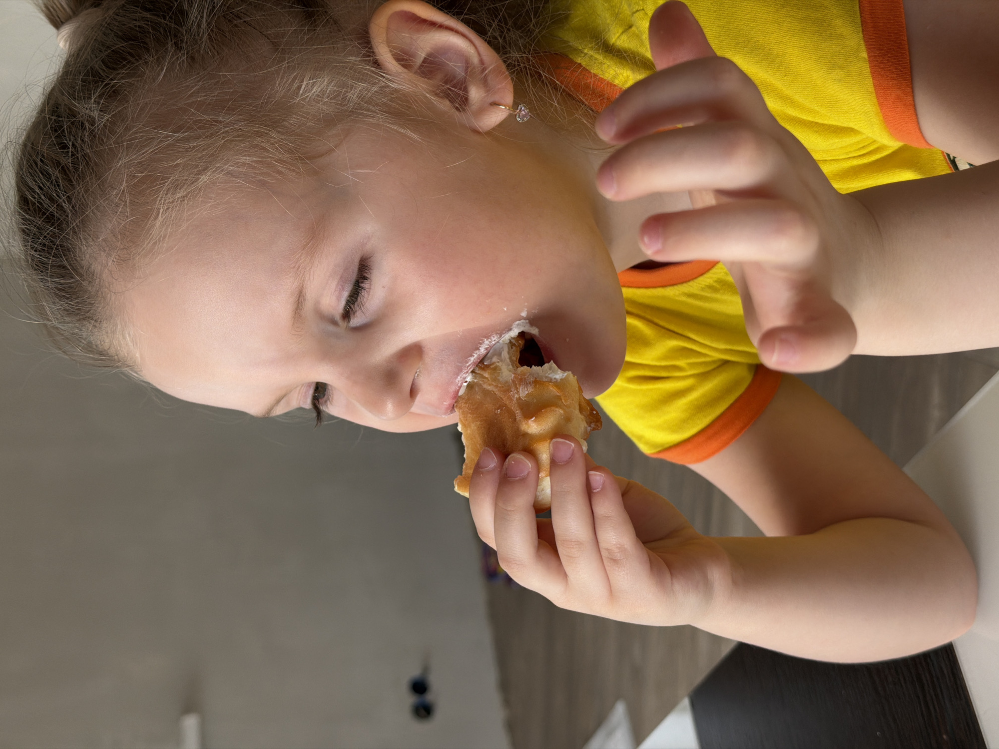 A young girl with blonde hair pulled back in a ponytail is shown in close-up, angled to the side, taking a bite of a cream-filled pastry. She wears a yellow shirt with orange trim. Her eyes are closed. She is holding the pastry in one hand, and the other hand is raised near her face. The background is a gray wall and a black and white surface.
There is no information in the image to suggest it was taken in Chaikovsky, Russia.