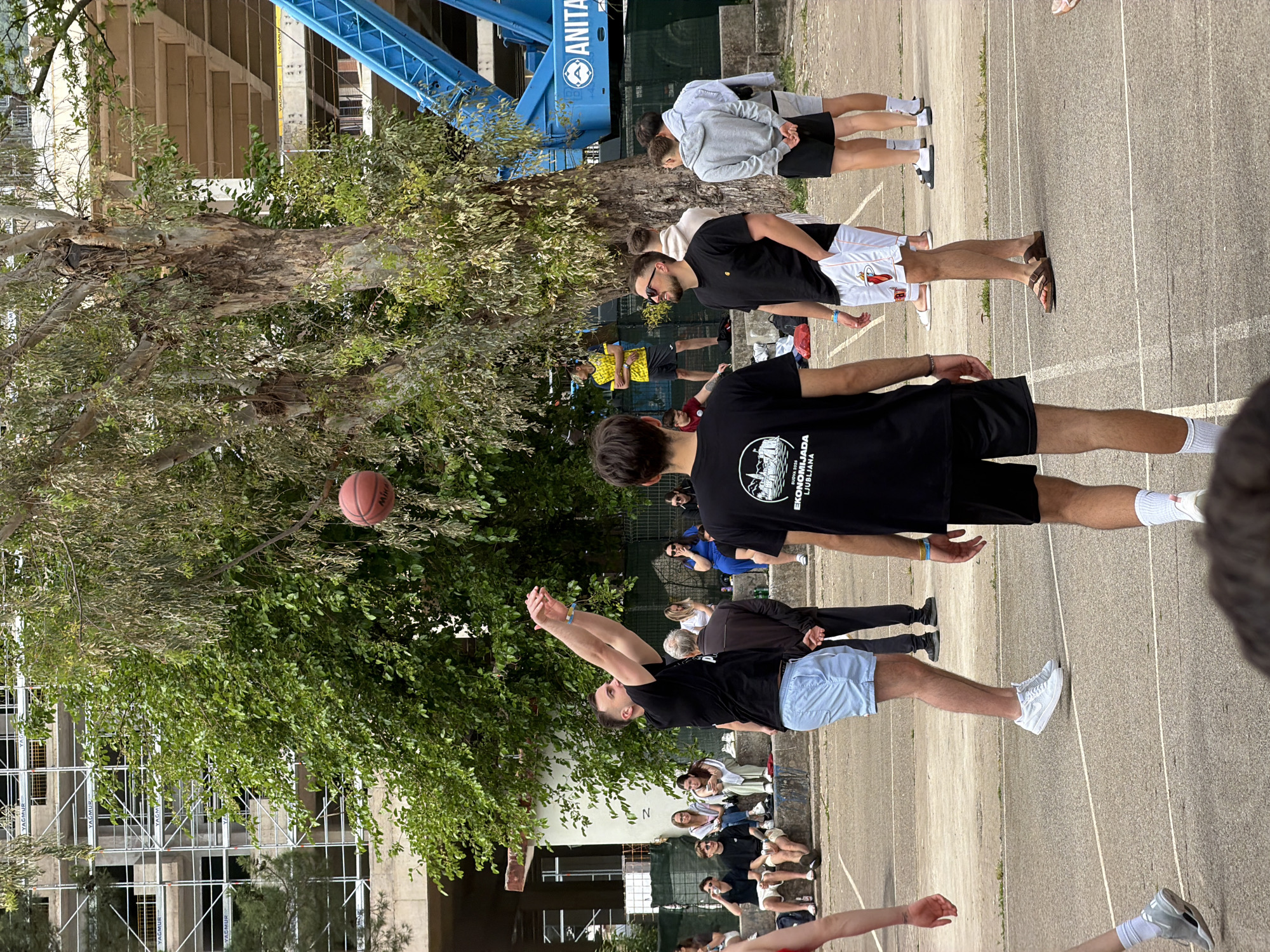 A photo depicts a group of people gathered on a concrete surface marked with lines, likely a sports court. One man in a black shirt and light blue shorts has his arms raised, as if he has just thrown a basketball, which is suspended in the air near a leafy tree. Other individuals are standing or sitting along the edge of the court, observing. Behind the court, there is a partially obscured building with scaffolding, and a blue support structure with the word "ANITA" visible. The overall setting suggests an outdoor location, possibly in a public park or recreational area. While the location cannot be absolutely determined from the image, the presence of the word "ANITA" could indicate that it is in Budva, Montenegro, but cannot be determined without absolute certainty.