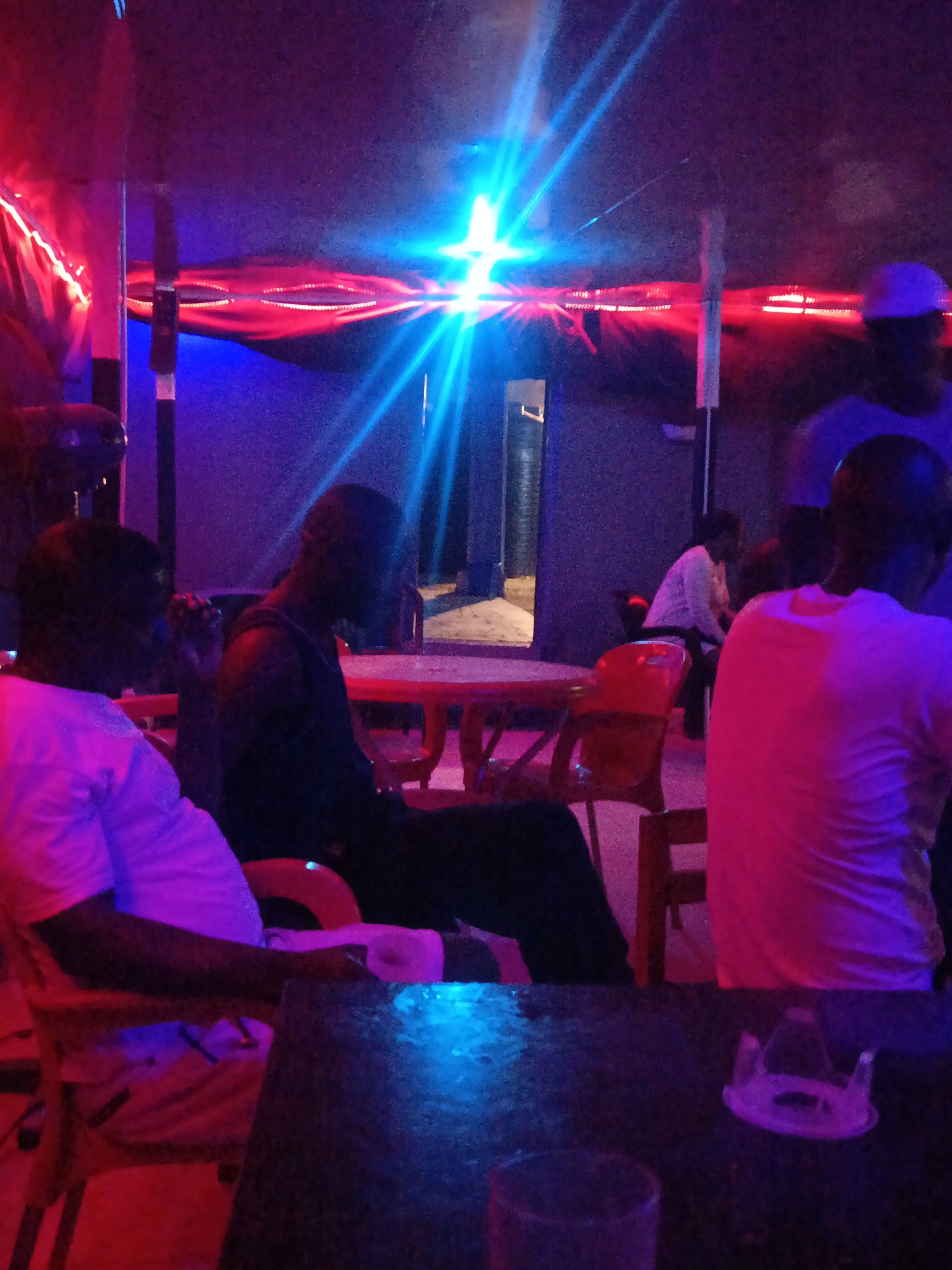 The image captures an indoor scene, likely a bar or lounge in Akwanga, Nigeria, characterized by dramatic and vibrant artificial lighting. The environment is dark, indicating it is nighttime.
Several individuals are present, most of whom appear to be men, seated at tables or standing. In the foreground on the left, a person in a light-colored short-sleeved shirt is seated at a table, facing away from the viewer. Next to them, another individual in a dark top is also seated. Further into the room, another person in a dark shirt is seated at a red table, viewed from the side. To the right, an individual wearing a light-colored shirt is seen from behind. In the background, another person in a white top is seated, and one person wearing a light-colored cap stands. The faces of the individuals are generally obscured by shadows and the intense lighting.
The setting features red plastic chairs and tables, suggesting a casual establishment. Bright red string lights or decorative fixtures are strung along the upper part of the room, adding to the club-like atmosphere. A powerful blue light source, possibly a spotlight, emits strong rays across the scene from the left, creating a striking visual contrast with the red lighting. The interplay of these vivid blue and red lights casts dramatic shadows and highlights throughout the room, creating a dynamic and lively ambiance. There is no visible text in the image.