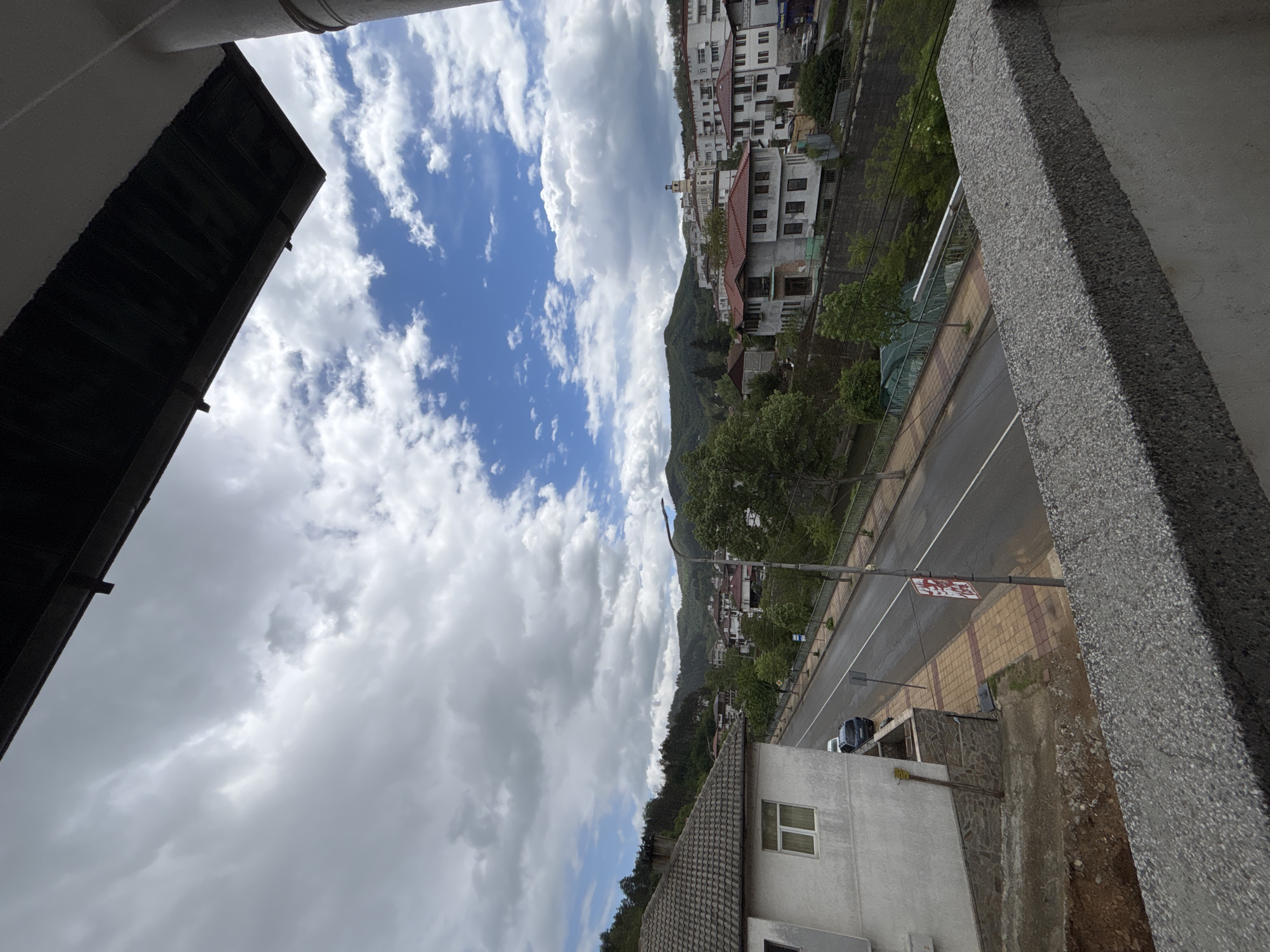 The image shows a view from an elevated position, possibly a balcony or high window, in Zlatograd, Bulgaria. The perspective is slightly tilted. The foreground consists of parts of the building the picture is taken from: a dark-colored shutter or door frame on the left and a light-gray concrete structural element on the right.
Beyond these foreground elements, a cityscape is visible. There are several buildings with varying architectural styles, ranging from residential houses to multi-story buildings, clustered along a mountain slope. The buildings are predominantly white or light-colored with red-tiled roofs.
A road runs parallel to the buildings and mountain slope. A gray car is parked on the side of the road, and a red sign is visible above the road. The road appears slightly wet, suggesting recent rain.
The background features a mountain partially covered with trees. The sky is filled with a mix of white and gray clouds, suggesting overcast weather conditions. Patches of blue sky are also visible.