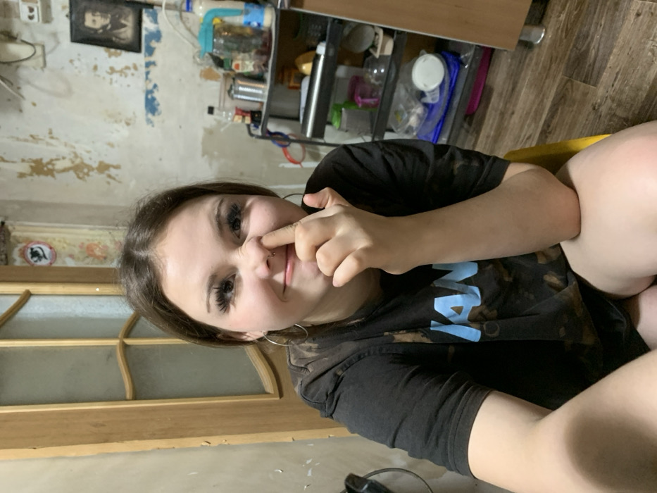 A woman with long eyelashes and a nose piercing sits indoors, playfully pinching her nose with her fingers. She wears a dark, tie-dye shirt and silver hoop earrings. A wooden door is behind her to the left. The wall has peeling paint, and a framed picture hangs on the wall above. Shelves containing various items are in the background on the right, positioned above a wooden floor.