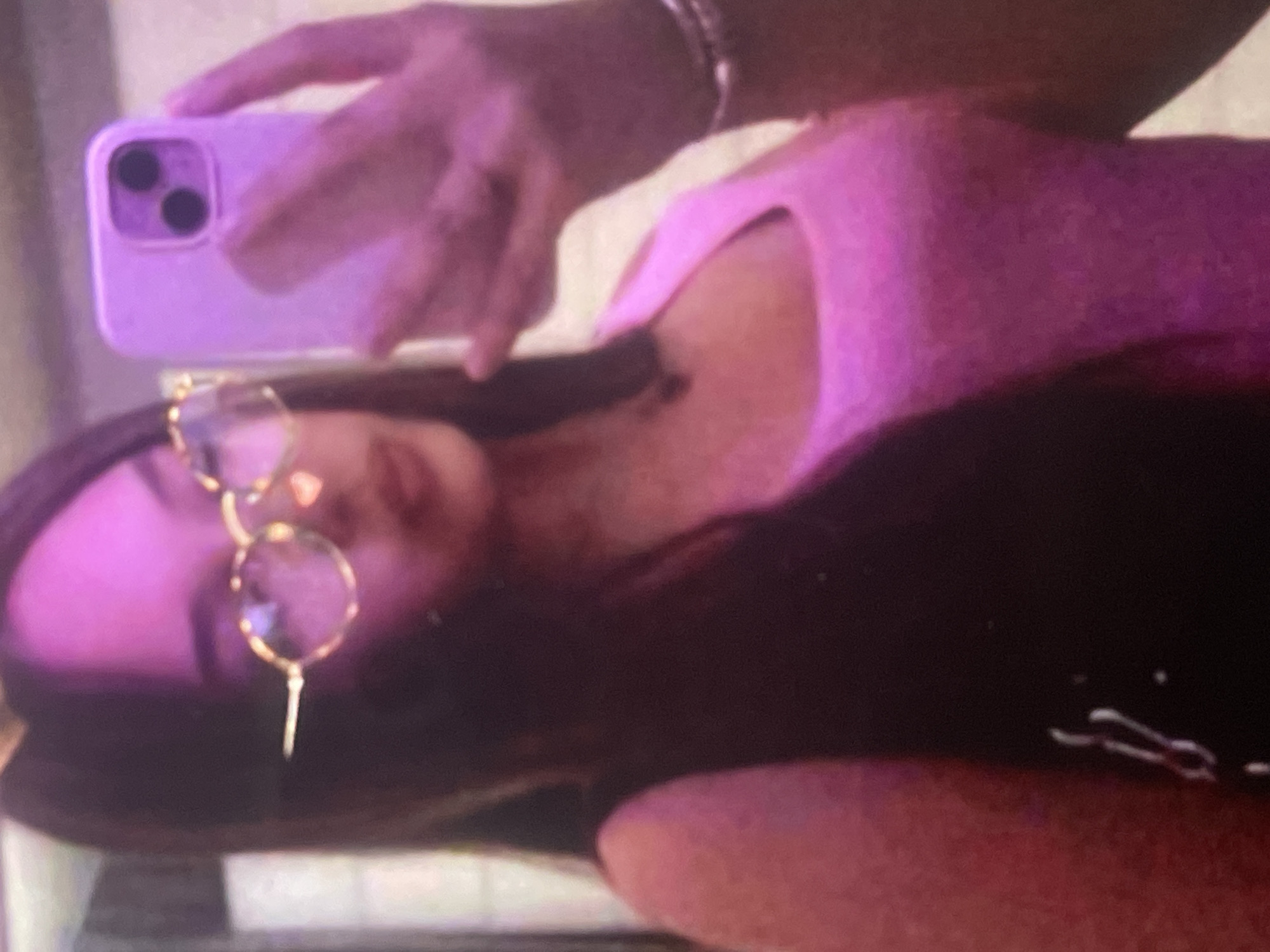 A mirror selfie taken indoors shows a person, likely a young woman, in Poltava, Ukraine. She has long dark hair and is wearing a light pink sleeveless top and distinctive round, gold-rimmed glasses. Her head is tilted, and she is looking towards the camera.
She is holding a light purple smartphone with a dual-camera system (two dark circular lenses) visible on its back. A hand with a thin bracelet is visible, holding the phone up to take the picture. The entire scene is bathed in a strong purple and pink artificial light, creating a vibrant and atmospheric feel, possibly suggesting evening or nighttime. No text is visible in the image. The person's expression appears relaxed, though details are slightly blurred.