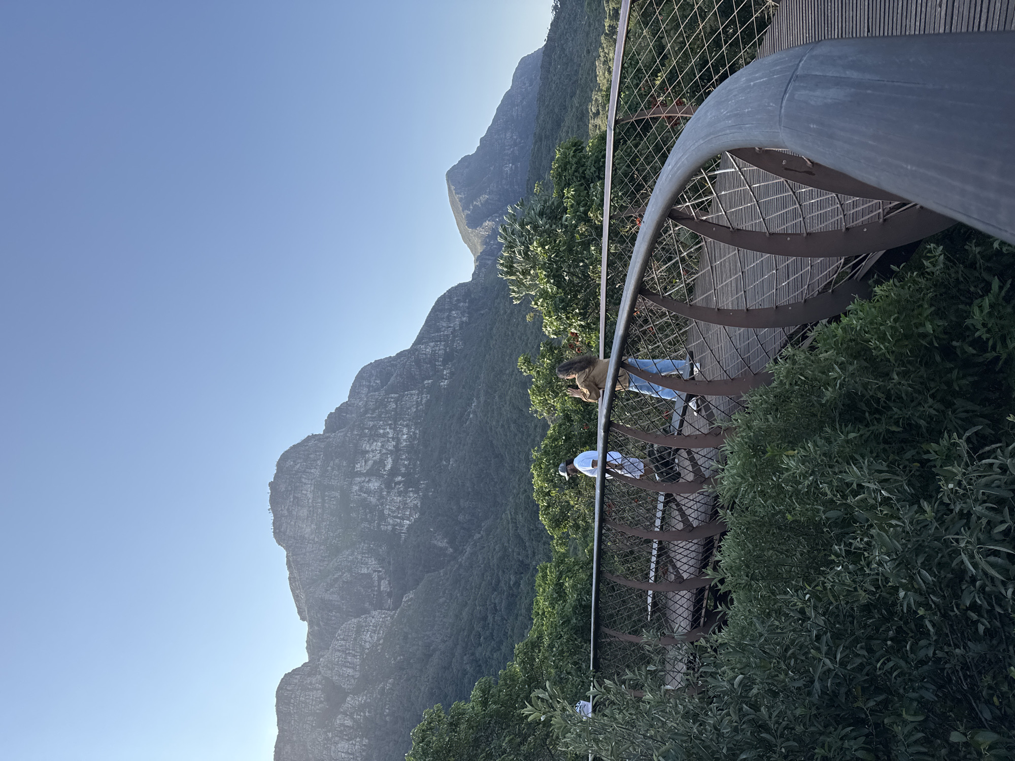 A curving elevated walkway with wooden planks and metal railings, partially obscured by lush green foliage in the foreground, is the central feature of the image. Two individuals are walking on the walkway; one, wearing a white t-shirt and shorts, is walking away from the viewer towards the left, and another, wearing a brown jacket and blue jeans, is facing towards the right and appears to be taking a photograph. In the background, a vast mountain range with rugged peaks and dense green forestation under a clear blue sky dominates the scene. This is a view of the Bo-Kaap neighborhood in Cape Town, South Africa, with the iconic Table Mountain in the distance. The lighting suggests it is either early morning or late afternoon.
