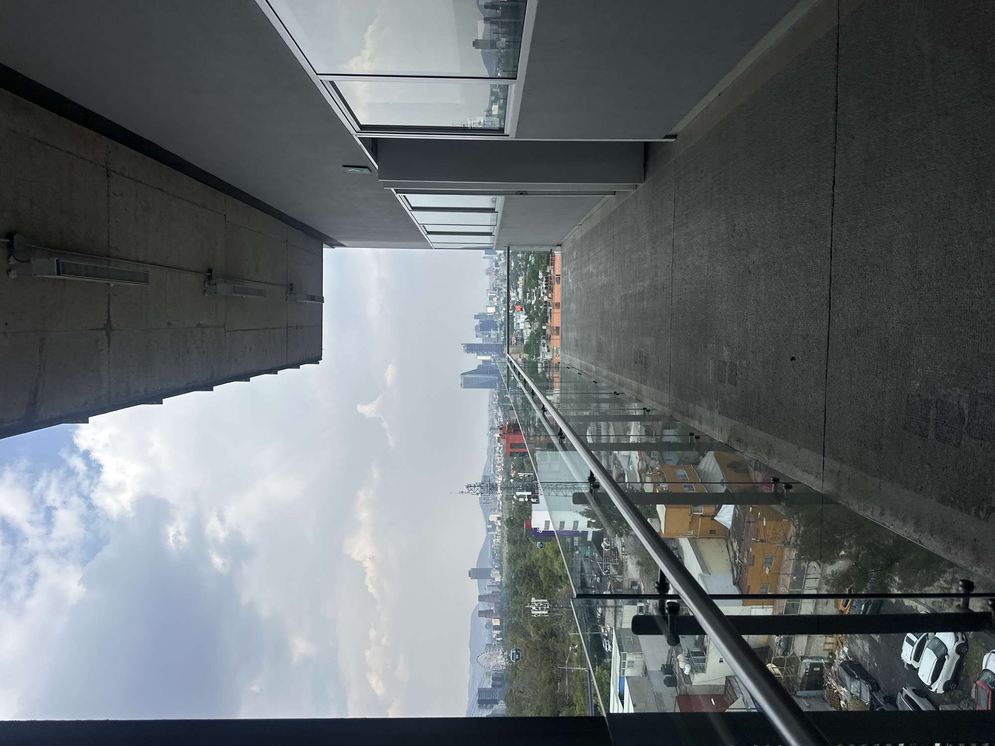The media file presents an upright view from a modern building's outdoor walkway or balcony, overlooking a sprawling city under an overcast sky.
In the foreground, to the left, is a tall, textured concrete wall with several metallic fixtures, possibly air vents or industrial lighting, mounted horizontally. Above this wall, a smooth grey ceiling or overhang extends. To the right, a contemporary glass and metal railing forms the barrier of the walkway, offering an unobstructed panorama. The concrete floor of the walkway is visible, leading towards the distant city.
The mid-ground reveals a dense urban landscape featuring a mix of architectural styles, including residential buildings with distinct color palettes (e.g., yellow, orange) and several taller, modern skyscrapers. Green areas with trees are interspersed within the urban fabric. A prominent Ferris wheel and communication towers are visible amidst the city buildings.
In the background, a range of mountains rises, partially obscured by the cloudy conditions. The sky is largely overcast, with varying shades of grey and white clouds, suggesting a cloudy daytime, possibly late afternoon, with diffused light. No direct sunlight or harsh shadows are present.
The scene depicts an expansive view of Mexico City, showcasing its diverse urban environment and natural surroundings. There are no people visible, and no discernible text is present in the image.