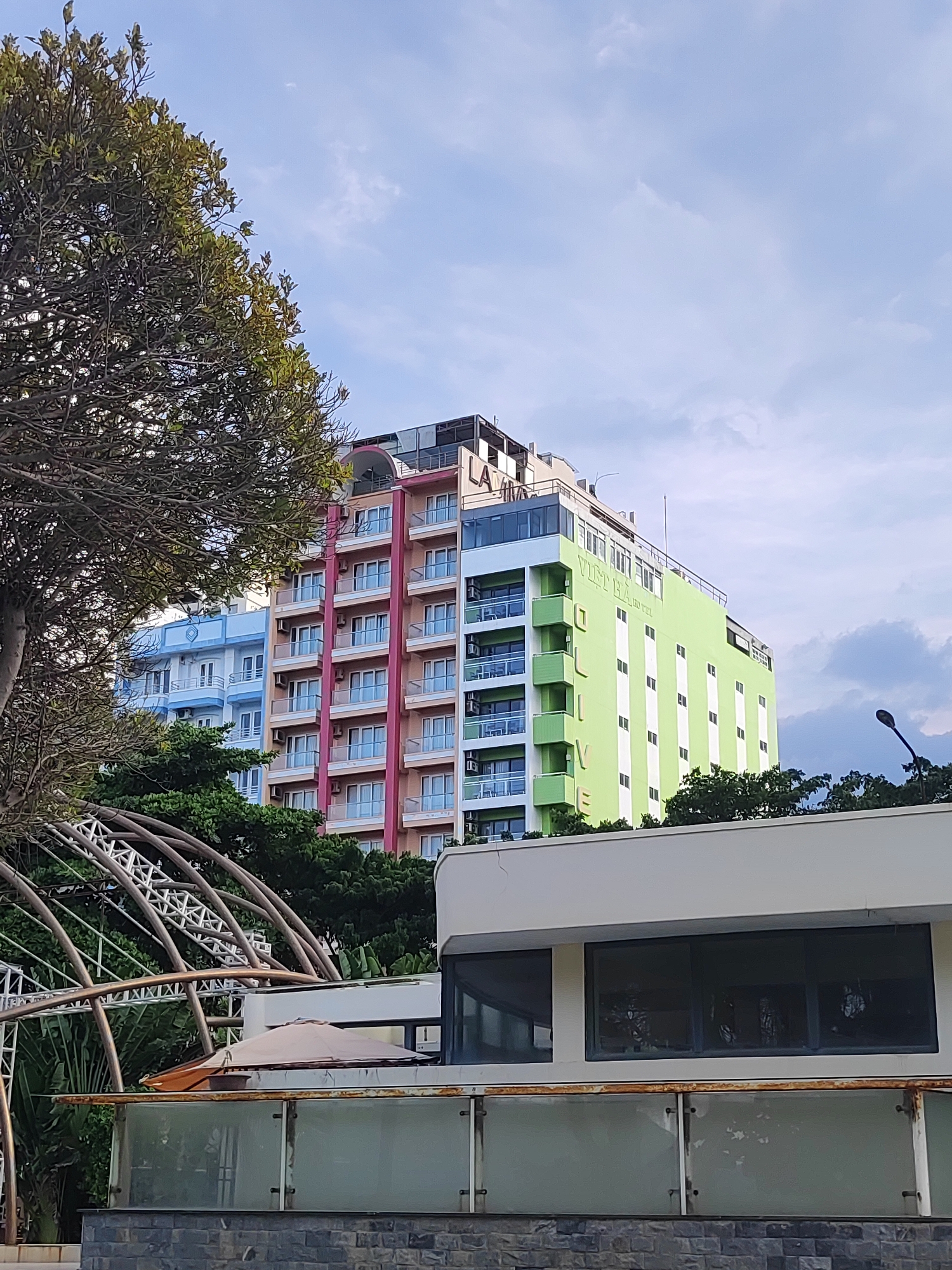 This media file captures an outdoor scene in Nha Trang Ward, Vietnam, likely during daylight hours given the bright sky and lighting. The focus is on a row of colorful buildings, appearing to be hotels or apartments, with distinct architectural styles. The leftmost building is painted light blue, followed by a building with pink and peach-colored facades, and the building closest to the right is a vibrant lime green. All buildings feature multiple stories with balconies and white-framed windows.
A large tree with dense, green foliage dominates the left side of the frame, partially obscuring the view of the buildings. In the foreground, there's a modern, semi-circular structure with a metallic frame, possibly part of a pavilion or walkway. Below this structure, a lighter-colored building with large windows and a glass-paneled railing suggests a commercial or hospitality establishment. A beige umbrella is visible on a balcony of this lower building.
The sky is partly cloudy, with soft white clouds against a pale blue backdrop. A streetlamp is visible on the right side of the frame, indicating an urban or semi-urban setting.
Visible text on the green building reads "VIET HA" and "HOTEL" in white lettering. The word "OLIVE" is also prominently displayed vertically in green letters on the side of the green building.
The overall impression is a bright, somewhat tropical, and modern coastal environment. No people are visible in the frame, and the scene appears to be peaceful and static. The combination of the colorful buildings, lush greenery, and architectural elements creates a visually engaging and distinctly Vietnamese landscape.