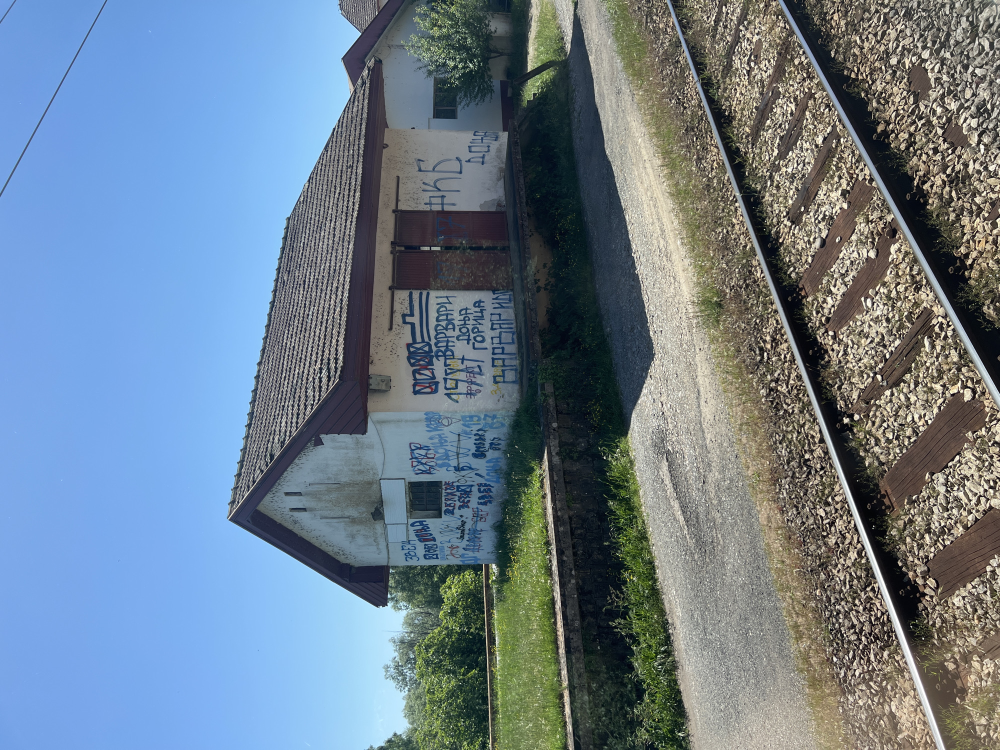 An exterior shot shows a two-story building covered in graffiti in Bistrice, Montenegro. The building, with a roof of terracotta tiles and white walls, stands adjacent to a railway track that runs alongside a dirt road. Graffiti is extensively spread on the building's walls. The sky is bright blue. Green foliage is visible at the base of the building. Overhead electric lines are visible in the upper left of the image.