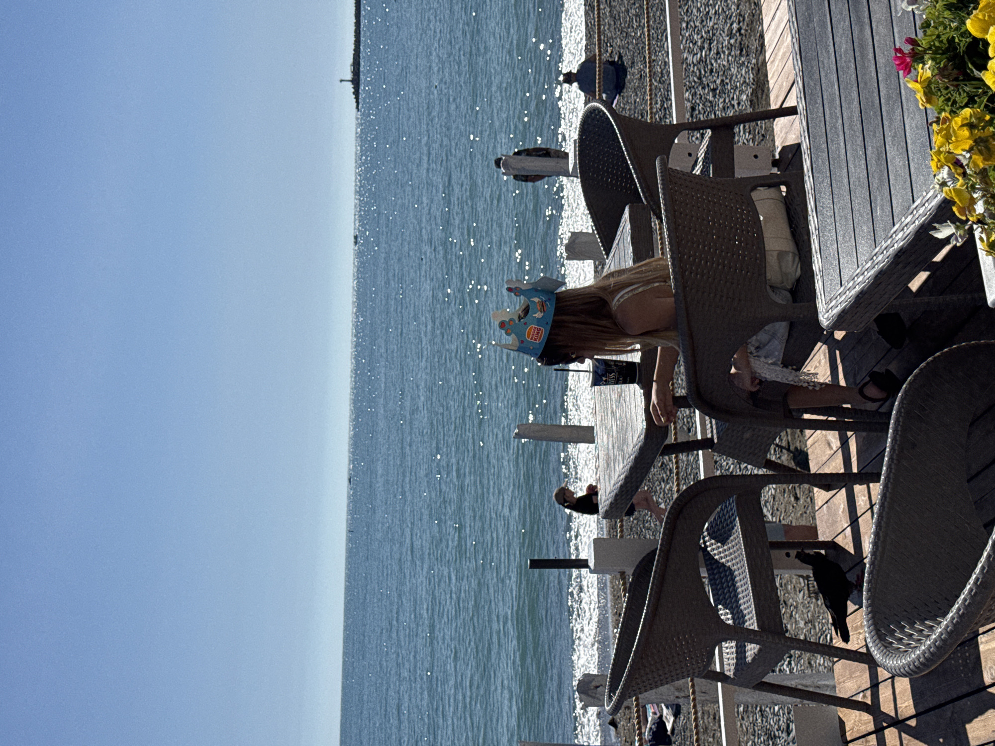 This image depicts an outdoor seaside cafe or restaurant in Sochi, Russia, offering a panoramic view of the Black Sea under a clear, sunny sky. The scene is captured from a wooden deck or pier, looking out towards the water.
In the foreground and mid-ground on the right, the cafe area is furnished with several modern wicker-style chairs and wooden slat tables. A decorative touch is added by a pot of vibrant yellow and some pink flowers adorning a table in the upper right. A black bird, resembling a crow or jackdaw, is perched on one of the empty chairs, indicating a relaxed atmosphere where wildlife is present.
A central figure, likely a young person, is seated at one of the tables. They have long blonde hair and wear a blue paper crown prominently featuring the "Burger King" logo, suggesting a casual outing or celebration. Other patrons are seated at tables further back on the deck, while several indistinct figures can be seen walking or standing on the pebbly beach and in the shallow waters along the shoreline, just beyond the cafe's seating area.
The vast expanse of the sea occupies the left and central portions of the frame, its surface shimmering with sunlight, reflecting the bright, light blue sky. A faint, slender structure, possibly a distant lighthouse or a section of a pier, is visible on the far horizon. The overall impression is one of a serene and leisurely day by the coast in a popular resort town.