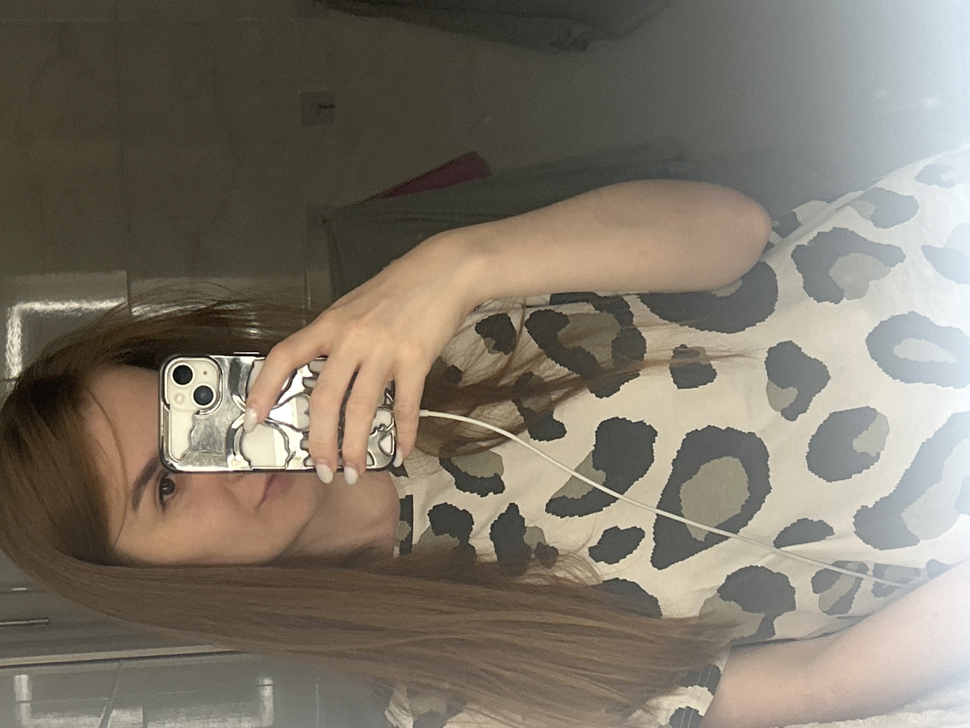 A young woman with long, auburn hair is taking a selfie in a mirror. She is wearing a tan and black leopard print t-shirt. Her right hand, with freshly manicured white nails, is holding up a silver phone with a skull-shaped case, obscuring her face. A white earphone cord is visible, running from the phone down her shirt. The background is dimly lit and appears to be a bathroom, with a mirror reflecting the scene and some hanging clothes in the distance. The image does not provide specific details to place it in the city of Нижние Муллы, Russia.