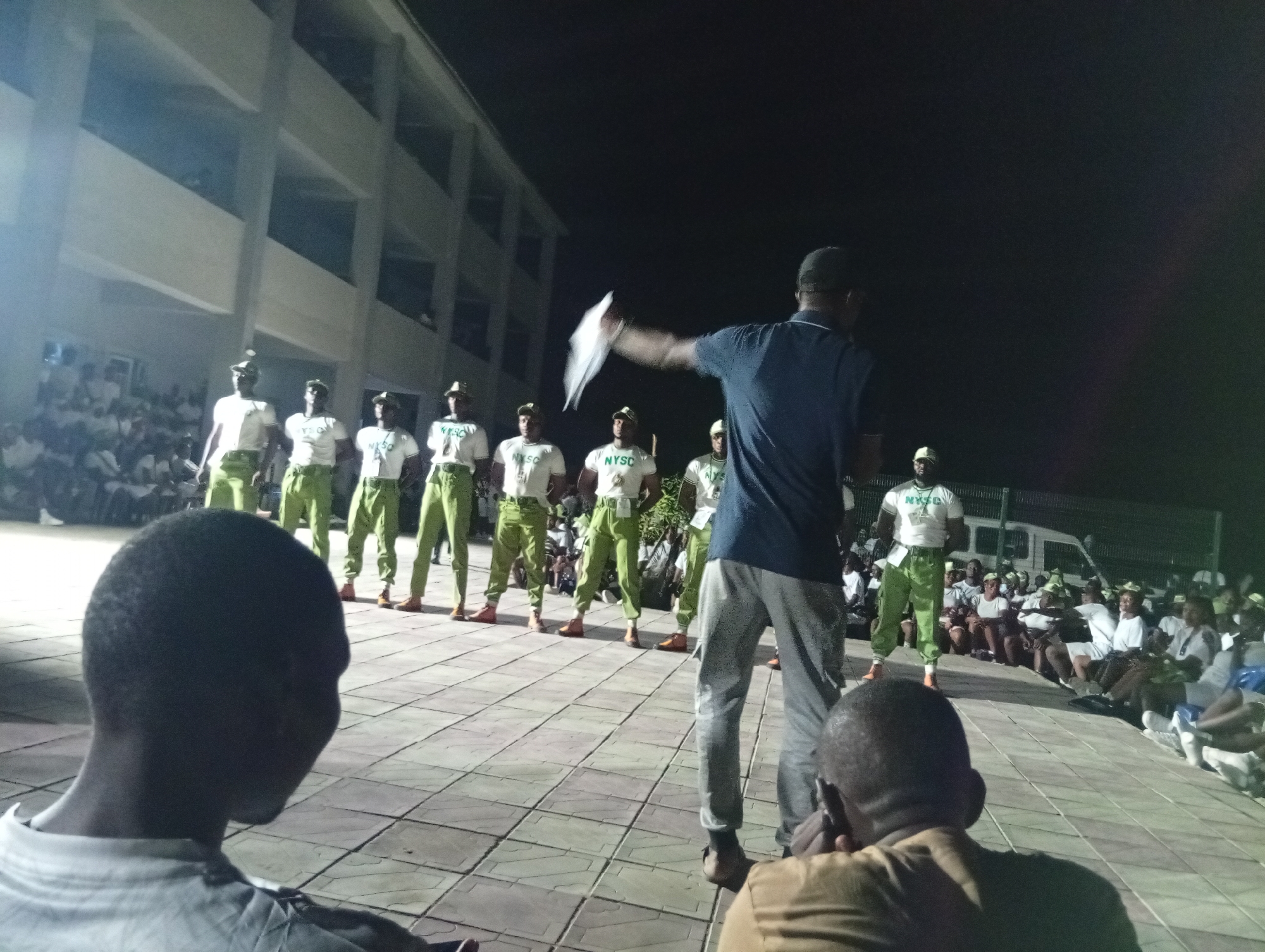 A nighttime image shows a performance or presentation in Maiduguri, Nigeria. A group of individuals wearing white T-shirts with "NYSC" printed on them and green trousers stand on a paved platform. A man in a blue shirt and grey trousers is facing them, holding a piece of white paper. Other people are seated, watching. A building is in the background.