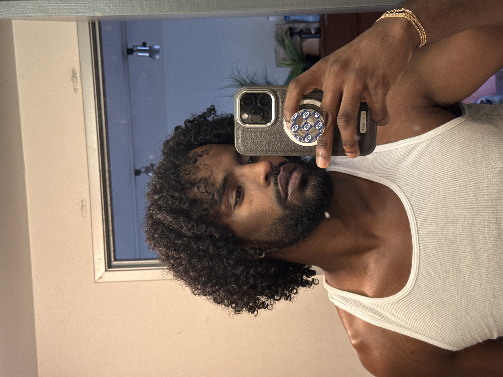 A selfie of a Black man in a white tank top, holding a phone with a patterned pop socket in front of a bathroom mirror. The mirror reflects a partial view of the bathroom, including a light fixture and possibly a plant. The man has curly hair and a beard. He is wearing a gold bracelet. The location is likely in a residential bathroom somewhere in Los Angeles, United States of America.