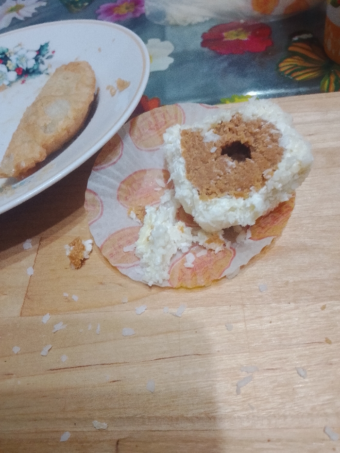 The scene is located in the city of Kaluga, Russia. The image displays a close-up, overhead view of a light-colored wooden surface. On the left, an oval white plate, adorned with a floral pattern, holds a single, elongated, golden-brown pastry that appears partially consumed, with a section missing from one end. To the right, a partially eaten round dessert rests within a white and orange patterned paper liner. This dessert features a broken white, flake-covered outer layer, exposing a dense, light brown core with a distinct dark cavity in its center. Scattered white, flake-like crumbs are visible on the wooden surface, particularly around the dessert. The background is blurred, revealing a multicolored tablecloth with various floral and abstract patterns. No individuals are present.