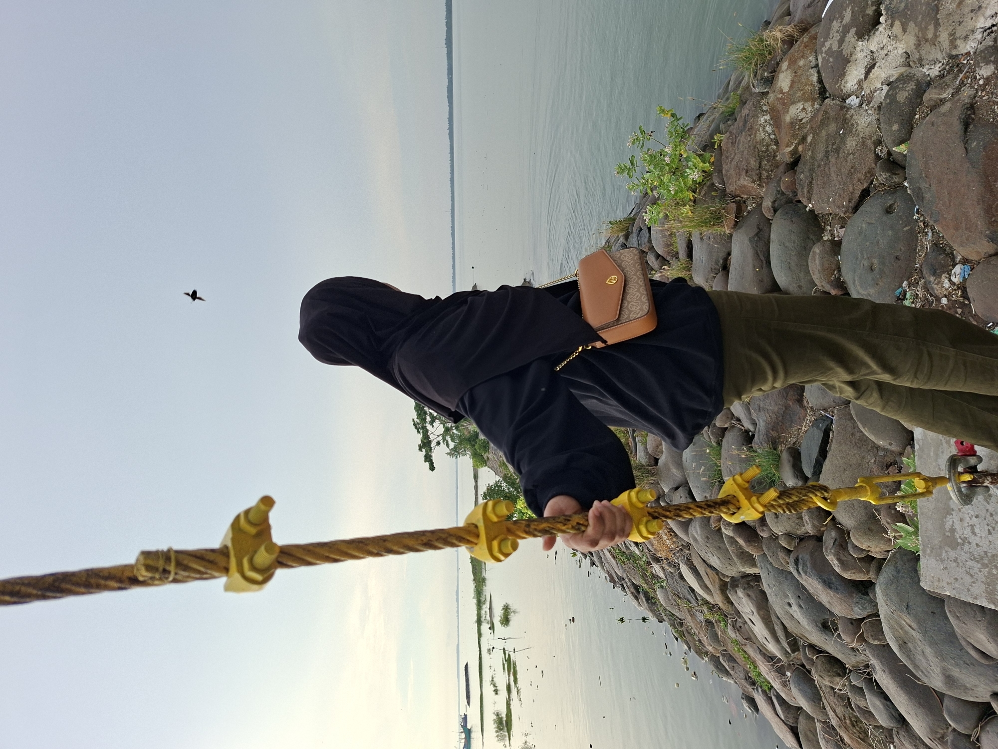 A person wearing a black hijab and black jacket stands on a rocky pier, looking out at a calm body of water. They are holding onto a thick yellow rope with yellow metal clamps. A small brown purse with a gold chain is slung across their body. The pier is constructed from large, rounded gray and brown rocks. Green vegetation grows along the edges of the pier and in patches on the rocks. In the distance, the water extends to the horizon, meeting a pale, overcast sky. A single black bird is visible in flight against the sky. A faint outline of land and possibly some structures can be seen on the far side of the water. A boat is also visible in the upper left portion of the image, further out on the water. The image appears to have been taken in the city of Lhokseumawe, Indonesia, near the coast.