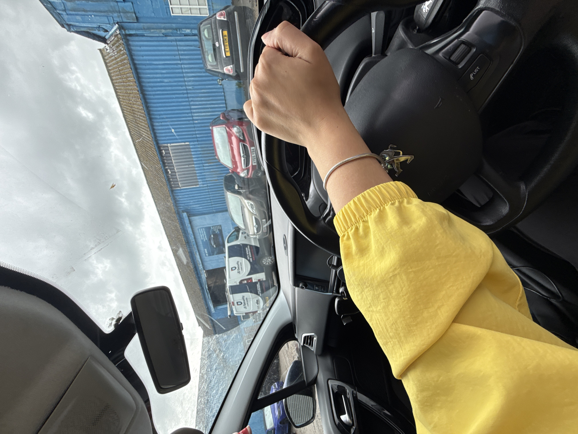 The image depicts a first-person perspective from the driver's seat of a car, looking out through the windshield. The scene is oriented as if looking upwards and to the right, with the driver's right arm and hand prominently featured on the left side, holding a black steering wheel.
The driver's right hand grasps the top of the steering wheel, which displays a Peugeot logo on the airbag cover. Controls are visible on the right spoke of the wheel, including a "LIST" button. A silver chain-link bracelet with a charm, possibly a rearing horse or lion, is worn on the driver's wrist. The driver's arm is clad in a bright yellow garment with a ruched or elasticated cuff. The car's interior, including parts of the dashboard and rearview mirror, is also visible.
Through the windshield, a cloudy, overcast sky is visible, suggesting a daytime setting without direct sunlight. Reflected and visible beyond the windshield is a blue corrugated metal building, typical of industrial or commercial architecture. Several vehicles are parked or moving in the background, including a black Fiat 500 with the UK license plate "ET11 UKB," a red car with the partially visible license plate "AY06 XYM," and two white vans. The vans are clearly marked with "JD Security" logos on their sides.
The setting appears to be an urban or industrial area, possibly a street or parking lot in Great Yarmouth, United Kingdom, as indicated by the license plates and the provided context. The activity seems to involve the person driving, waiting, or being stationary in the car. No specific emotions can be discerned.