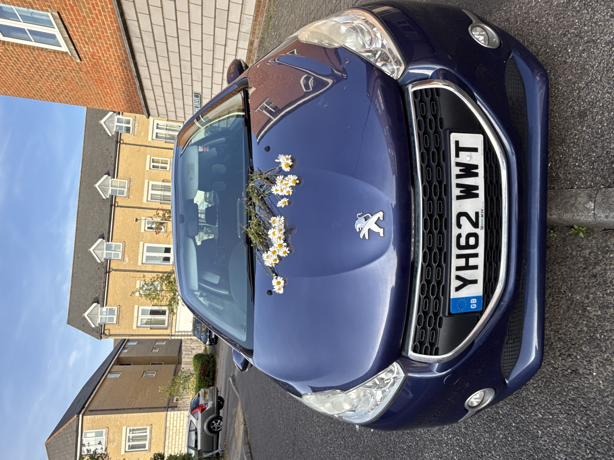 The image shows the front of a blue Peugeot car parked on a street in a residential area. The car has a license plate reading "YH62 WWT". A bunch of daisies are resting on the hood of the car, near the windshield. Behind the car are several two-story residential buildings made of yellow brick with dark roofs, along with a brick building that has some windows. A silver car is also visible parked in the background. The sky is blue with some clouds. There are green plants and a sidewalk bordering the street. The image is rotated clockwise.