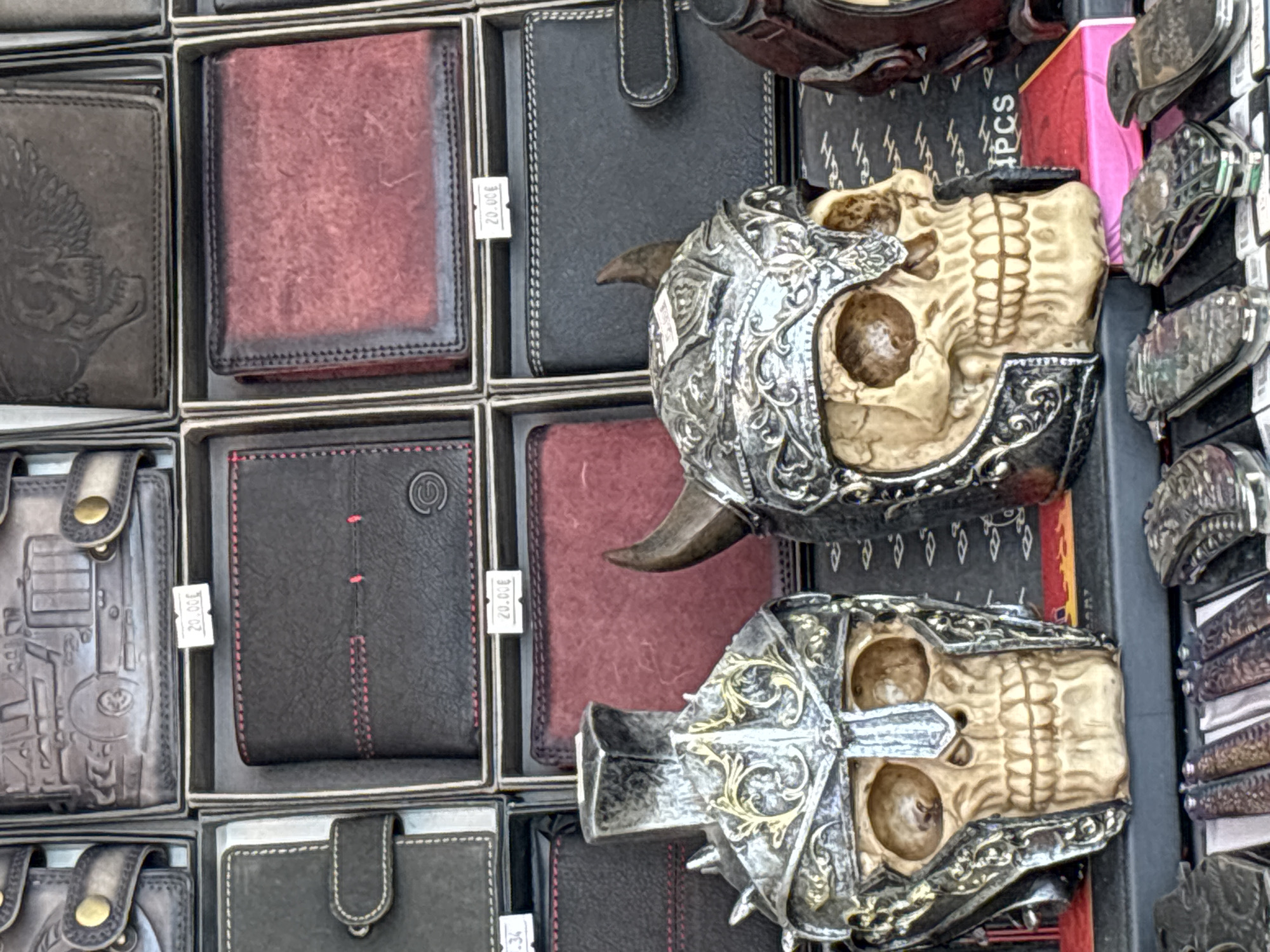 The image shows a display of wallets and skull-themed decorative items, likely in an outdoor market stall in Nesebar, Bulgaria. In the foreground, two skull figurines with elaborate metal helmets are prominently displayed. The skull on the left wears a helmet with spikes and golden accents, while the skull on the right has horns and a more ornate metal headdress. Behind these figures, a variety of wallets are arranged in tiered displays. Some wallets are black with decorative stitching, one is a deep red color, and others appear to have embossed designs, including a truck and a skull. Price tags, some indicating "20.00€", are visible on some of the wallet displays. The background suggests a bustling market environment, with other goods and stalls possibly present just out of frame. The overall lighting suggests daylight.
