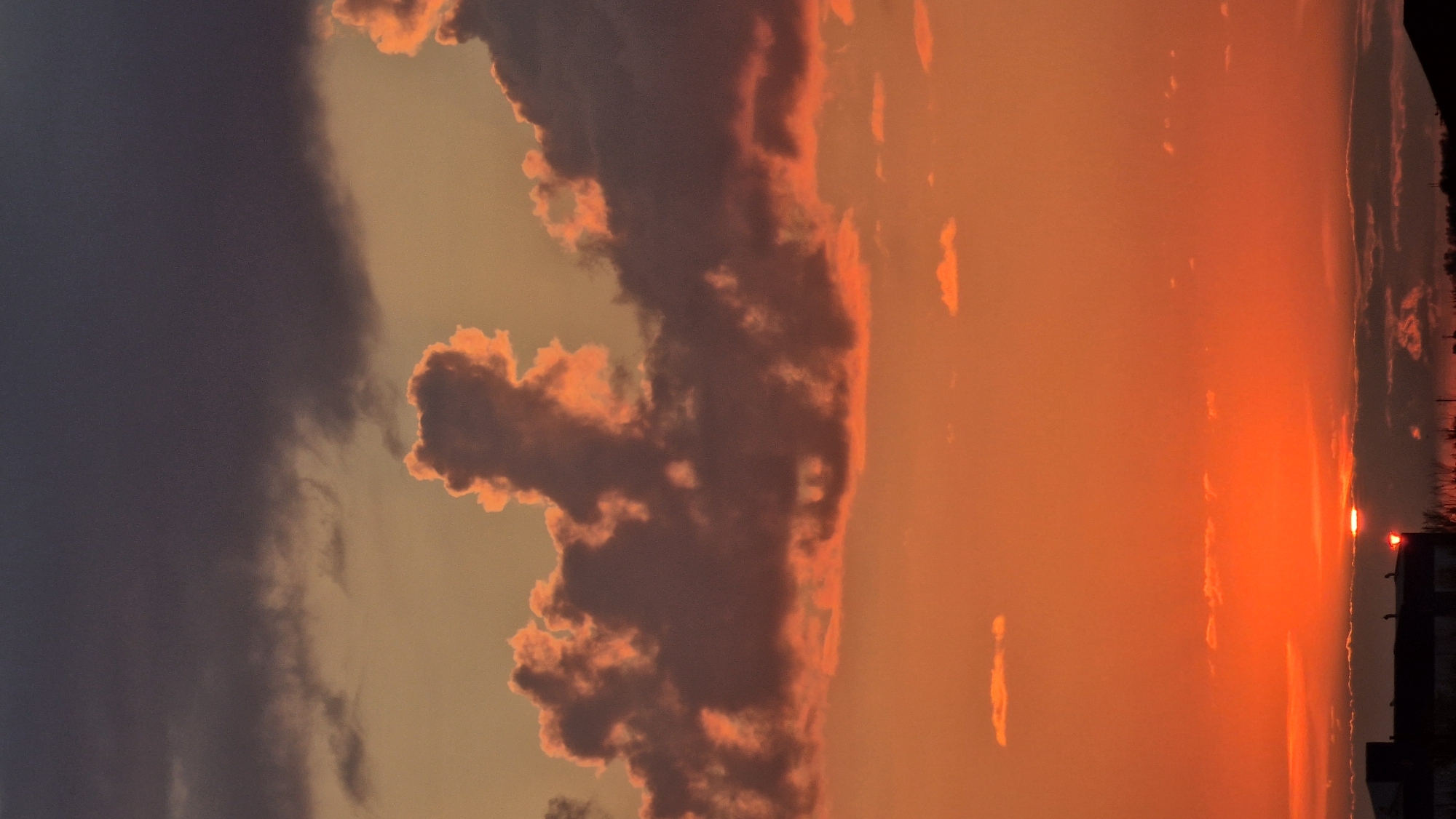 The image shows a sunset over the city of Kostanay, Kazakhstan. The sky is filled with a gradient of orange and yellow hues, with dark gray clouds towards the top and fiery orange clouds closer to the horizon. The sun is a small, bright point peeking through the clouds near the bottom center of the frame. Silhouetted against the vibrant sky are the rooftops of several buildings, including what appears to be a darker, larger structure on the left. The overall scene depicts a dramatic and colorful end to the day.