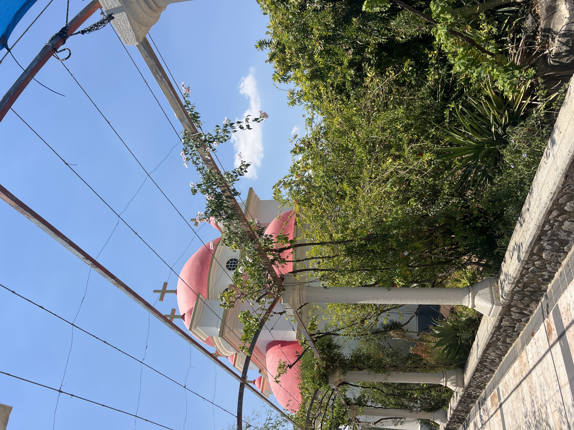 The image shows a building with red domes and crosses on top, likely a church, situated in a sunny outdoor environment. The building is partially obscured by a pergola with vines and flowering plants, supported by white columns. In the foreground, a stone wall runs along a paved walkway. Lush green vegetation fills the middle and background. The sky is clear blue with a few scattered clouds. The location is identified as Emek HaYarden Regional Council, Israel.