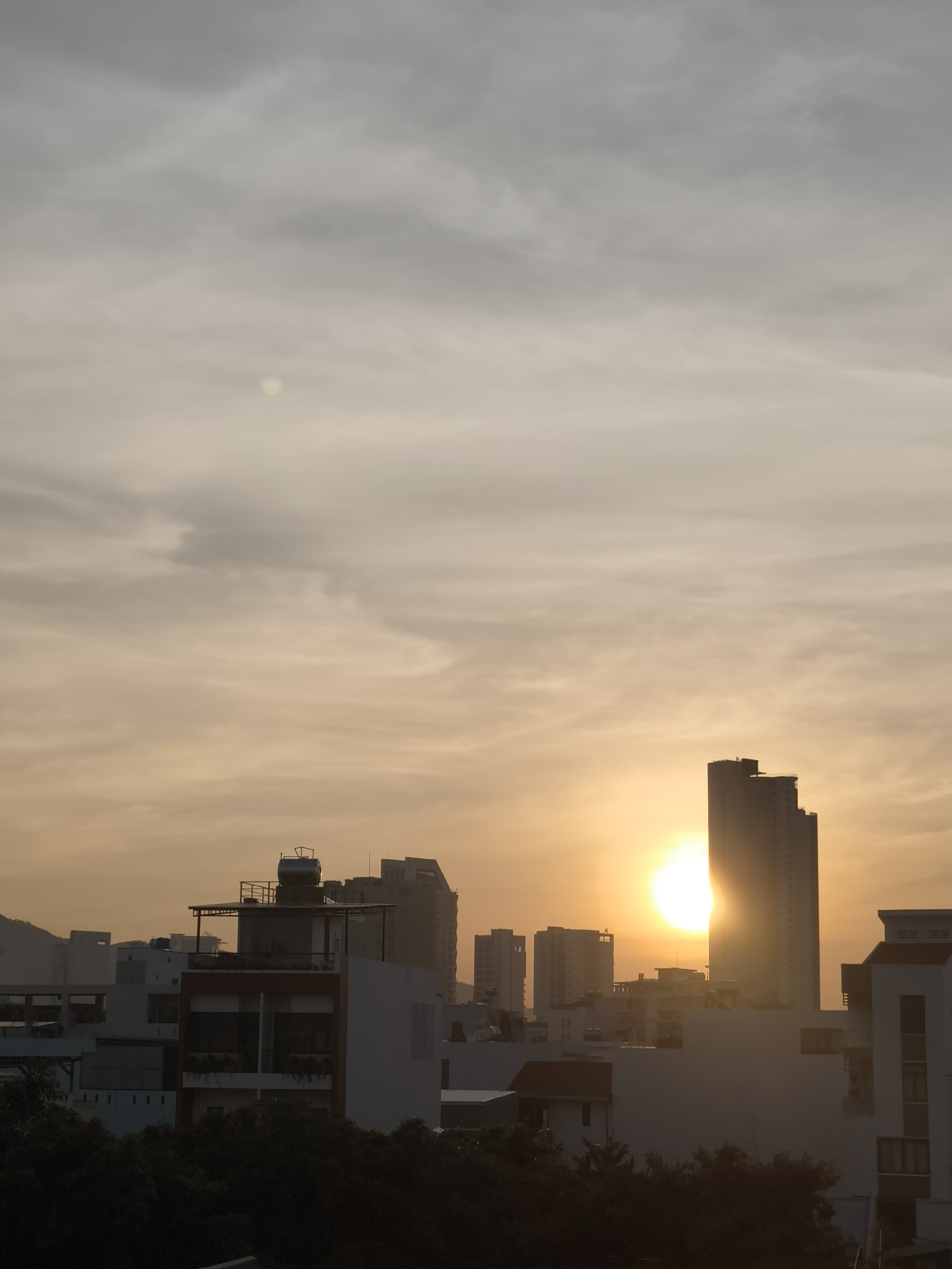 This is a vertical, eye-level, full shot of a city skyline at sunset in Bac Nha Trang Ward, Vietnam. The sun is a bright, orb-like presence in the middle right of the frame, partially obscured by a tall, rectangular building. The sky is a mix of light gray and muted orange clouds. Below the sky, several buildings of varying heights are silhouetted against the sunset. In the foreground, dark green trees form a dense line across the bottom of the image. The overall atmosphere is serene and captures a quiet moment in the city.