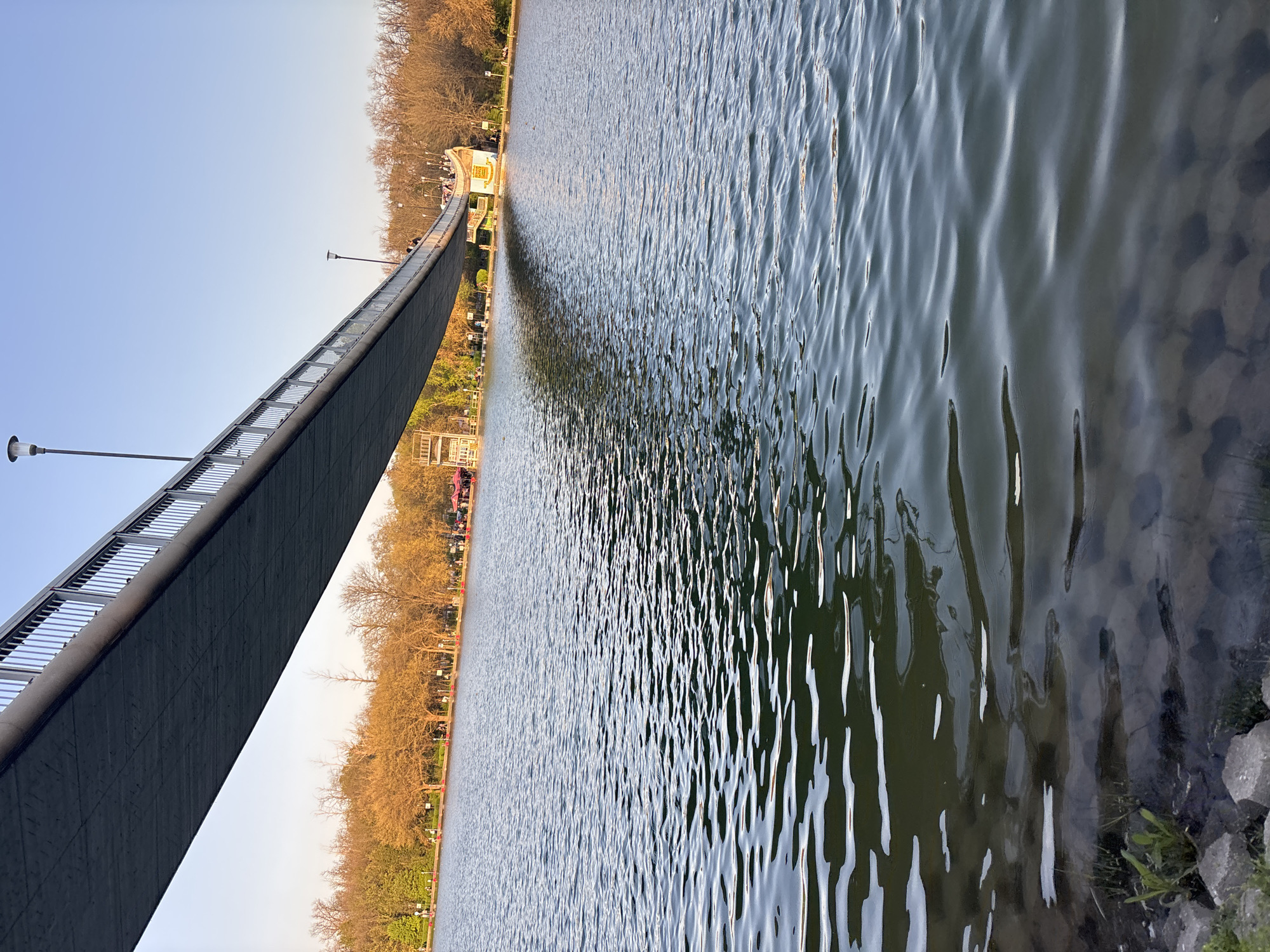 The image shows a long pedestrian bridge over a lake in Plovdiv, Bulgaria. The bridge, with metal railings and lampposts, stretches from the lower left to the center of the frame. People can be seen walking on the bridge. A line of trees lines the lakeshore on one side, behind which are various structures and a walkway. The lake fills the majority of the right side of the frame. The water's surface is rippled. The sky is clear and blue.