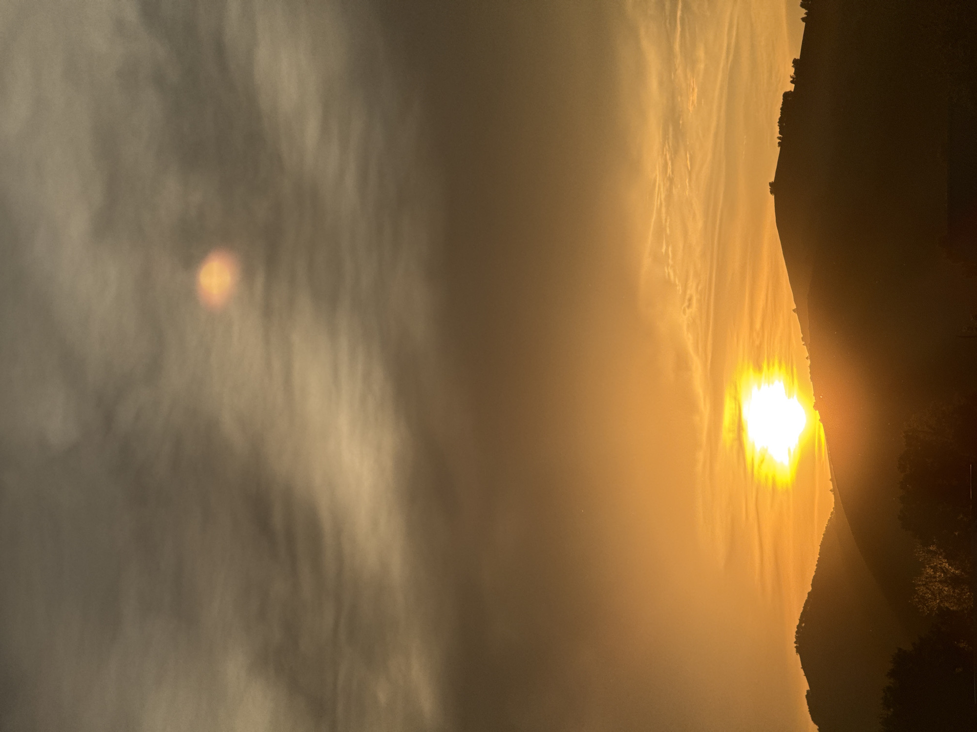 This is a vertical, eye-level, full-shot photograph of a sunset over a hilly landscape. The sky is dominated by shades of orange and brown, with clouds creating a textured, layered effect. The sun, a bright, white orb, is positioned in the lower half of the frame, casting a golden glow that illuminates the clouds and the upper portions of the hills. The hills themselves are silhouetted against the bright sky, with dark trees visible at the bottom of the frame. The location is specified as Soloneshnoye, Russia, though no specific landmarks are visible that would pinpoint this exact location within the city. There are no people or animals present in the image. The overall atmosphere is serene and dramatic due to the intense colors of the sunset.